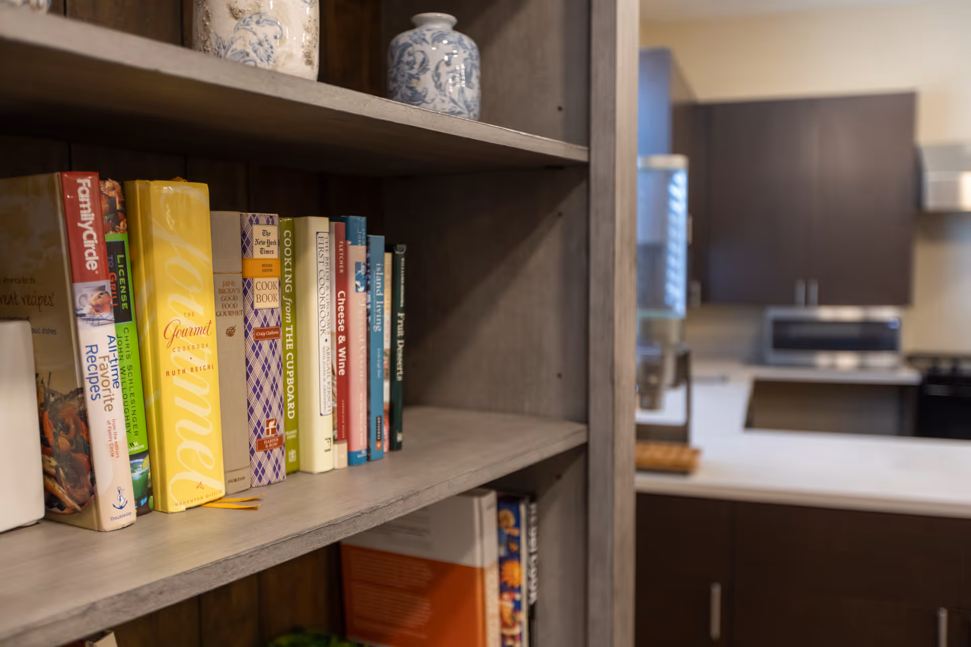 Wooden shelf filled with cookbooks in the foreground with a modern kitchen and countertop blurred in the background.