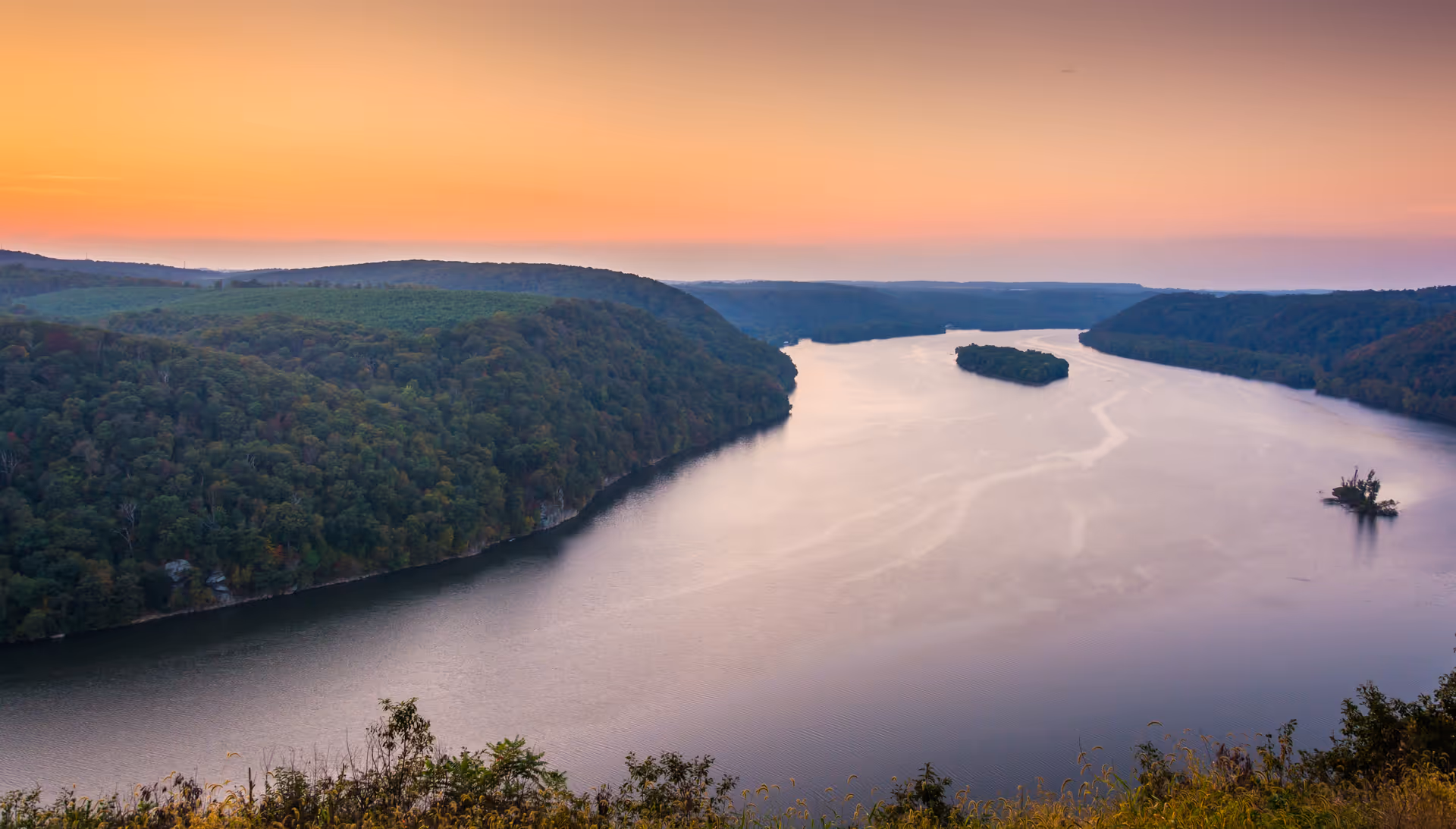 A wide river flowing between forested hills under a clear sky at sunset, with the horizon glowing in shades of orange and pink.