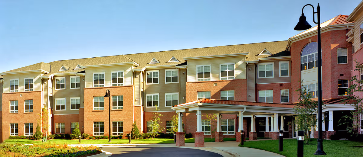 Exterior view of a large three-story senior living facility building with a combination of brick and beige siding. The building features multiple windows, a covered entrance with white columns, and a well-maintained lawn with small trees and shrubs. A black street lamp is visible near the entrance under a clear blue sky.