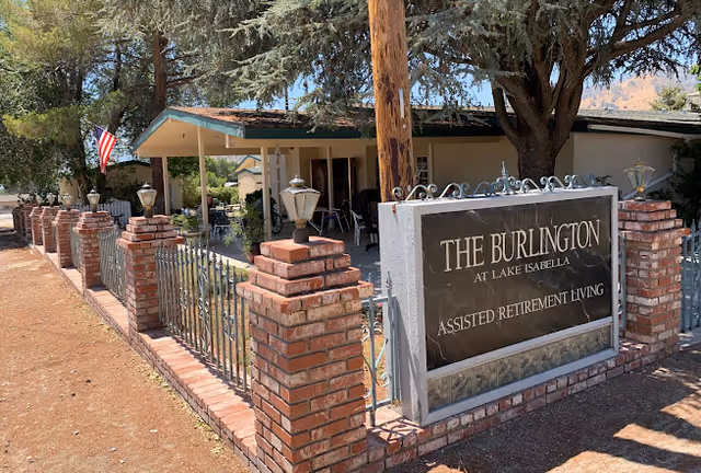Exterior view of The Burlington Assisted Living facility showing a brick and metal fence with lamp posts, a sign reading 'THE BURLINGTON AT LAKE ISABELLA ASSISTED RETIREMENT LIVING', and a building with a covered porch partially visible behind trees.