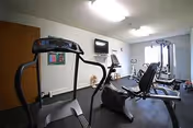 A fitness room with exercise equipment including a treadmill, stationary bike, and weight machines. The room has white walls, a wall-mounted TV, a clock, and a window letting in natural light.