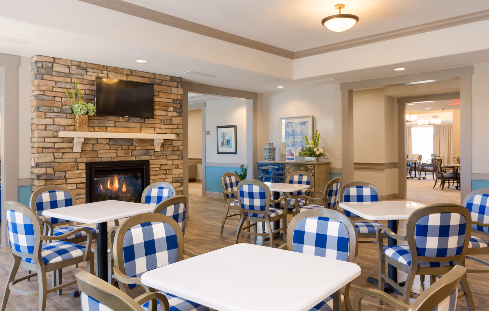 A cozy dining area in a senior living facility featuring white square tables surrounded by chairs with blue and white checkered cushions. A stone fireplace with a mounted flat-screen TV above it is on one wall. The room has wood flooring, soft lighting, and decorative plants and artwork. Adjacent rooms with additional seating are visible through open doorways.
