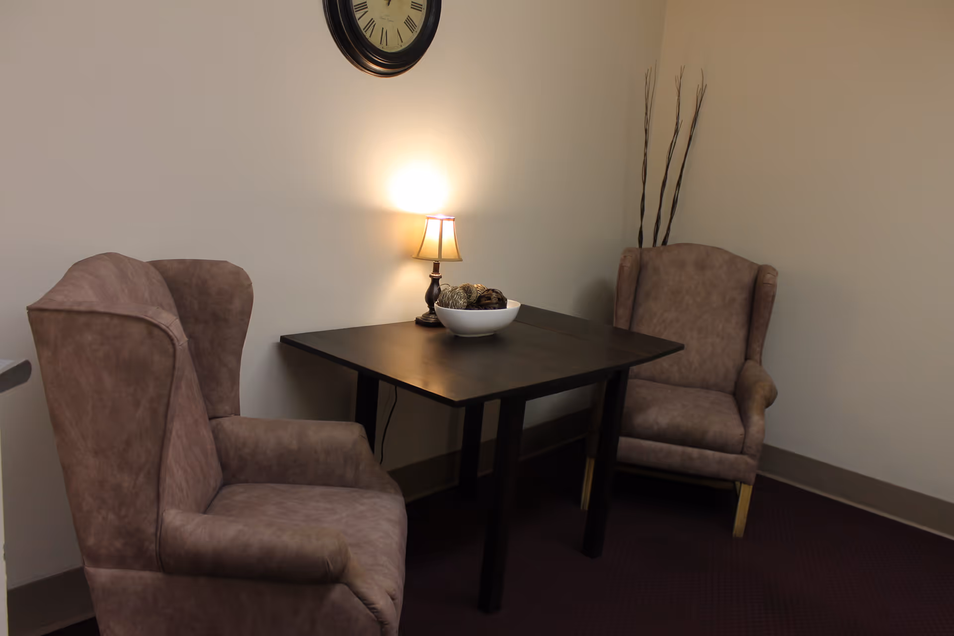 A small corner seating area with two brown upholstered armchairs facing a dark wooden square table. On the table is a small lamp emitting warm light and a white bowl with decorative items. A round wall clock hangs above the table, and tall decorative sticks are placed in the corner against the beige walls.