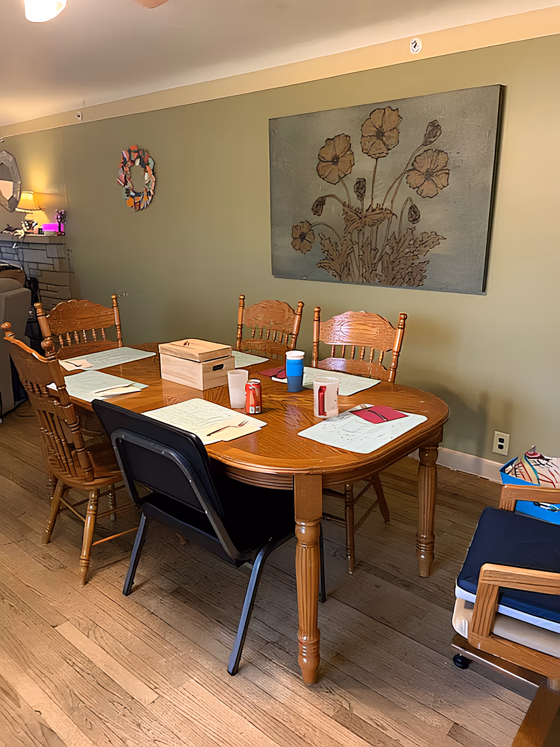 A wooden dining table set with six chairs, five wooden and one black plastic, placed on a wooden floor. The table has placemats, cups, napkins, and a wooden box on it. The background wall is light green with a large floral artwork hanging on it. There is a fireplace and a lamp visible in the background to the left.
