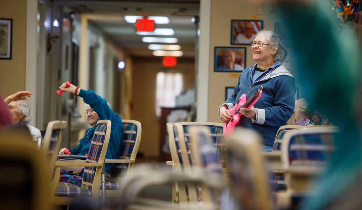 Older adults in a senior living community room doing seated exercises while a smiling woman holds a pink toy guitar.