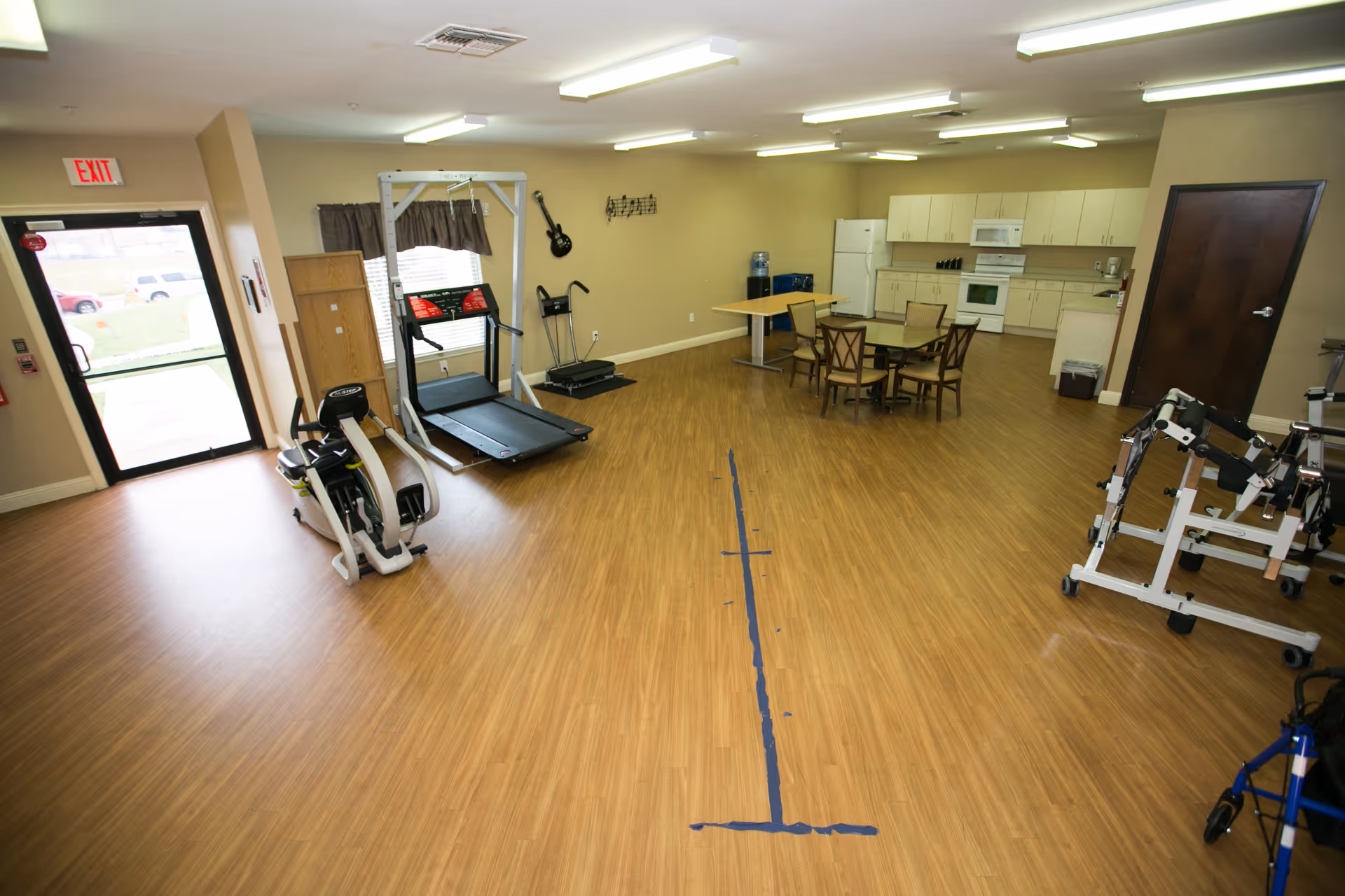 A spacious room with wooden flooring featuring exercise equipment including a treadmill, rowing machine, and other rehabilitation devices. There is a small dining area with a round table and chairs near a kitchenette with white cabinets, a refrigerator, microwave, and stove. The room has beige walls, fluorescent ceiling lights, and a glass door leading outside.