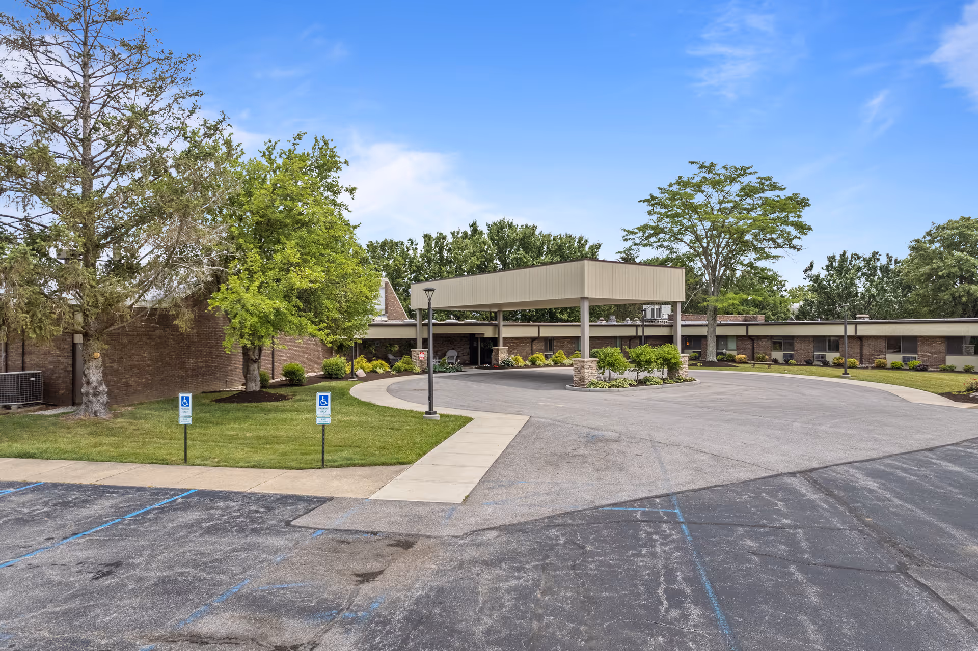 Front entrance of a single-story nursing facility with a covered drop-off, circular driveway, parking area, and landscaped greenery.