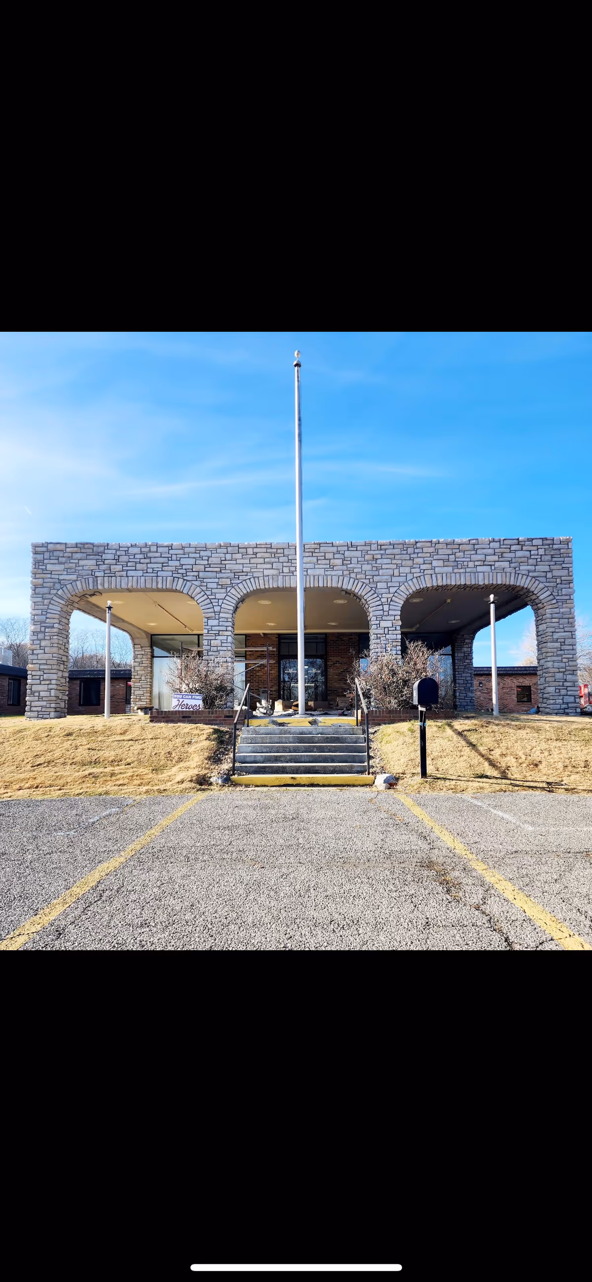 Front exterior view of a building with a stone facade featuring three large arches. There are steps leading up to the entrance, a flagpole in the center, and a mailbox on the right side. The sky is clear and blue.