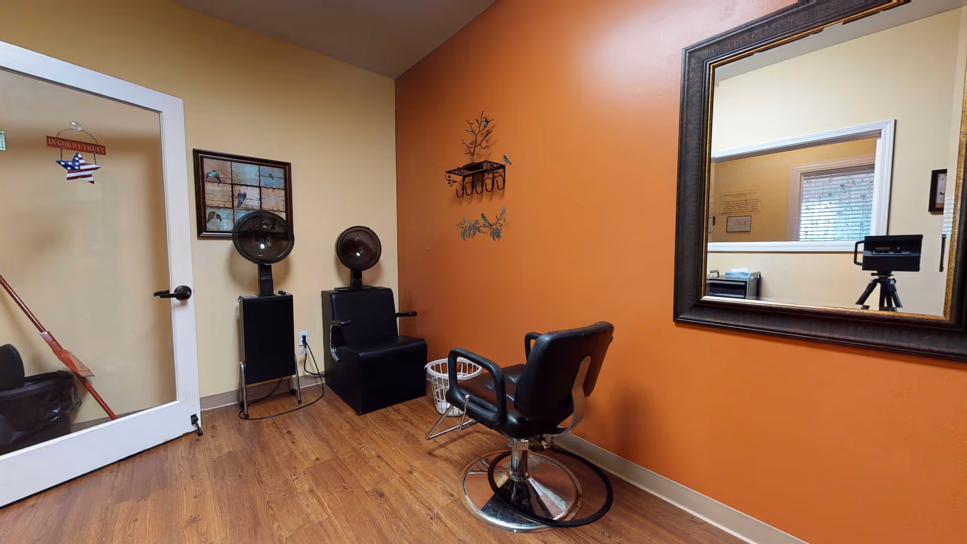 Interior view of a small salon or hair styling area with a black salon chair in front of a large framed mirror on an orange wall. Two black hair dryer chairs are positioned against a beige wall next to a door with a glass panel. The floor is wooden, and there is a small white basket on the floor near the salon chair.