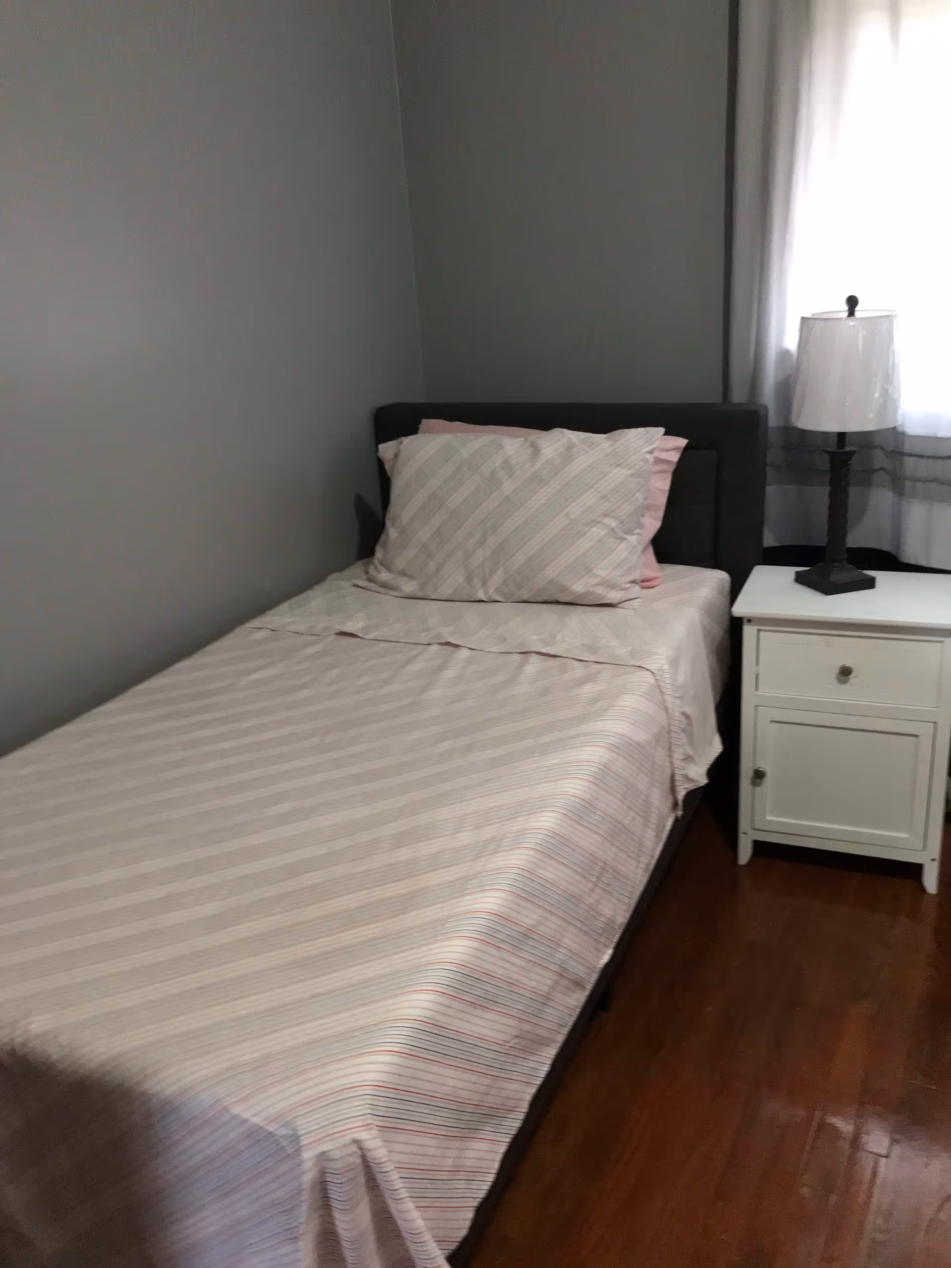 A small bedroom with a single bed covered in light-colored striped bedding and two pillows. Next to the bed is a white nightstand with a drawer and cabinet, topped with a lamp. The room has gray walls and a window with white curtains allowing natural light in.