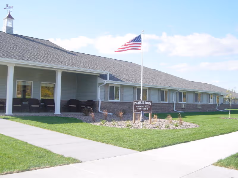 Single-story Prairie Home building front with a covered porch, American flag, and sign on a landscaped lawn.