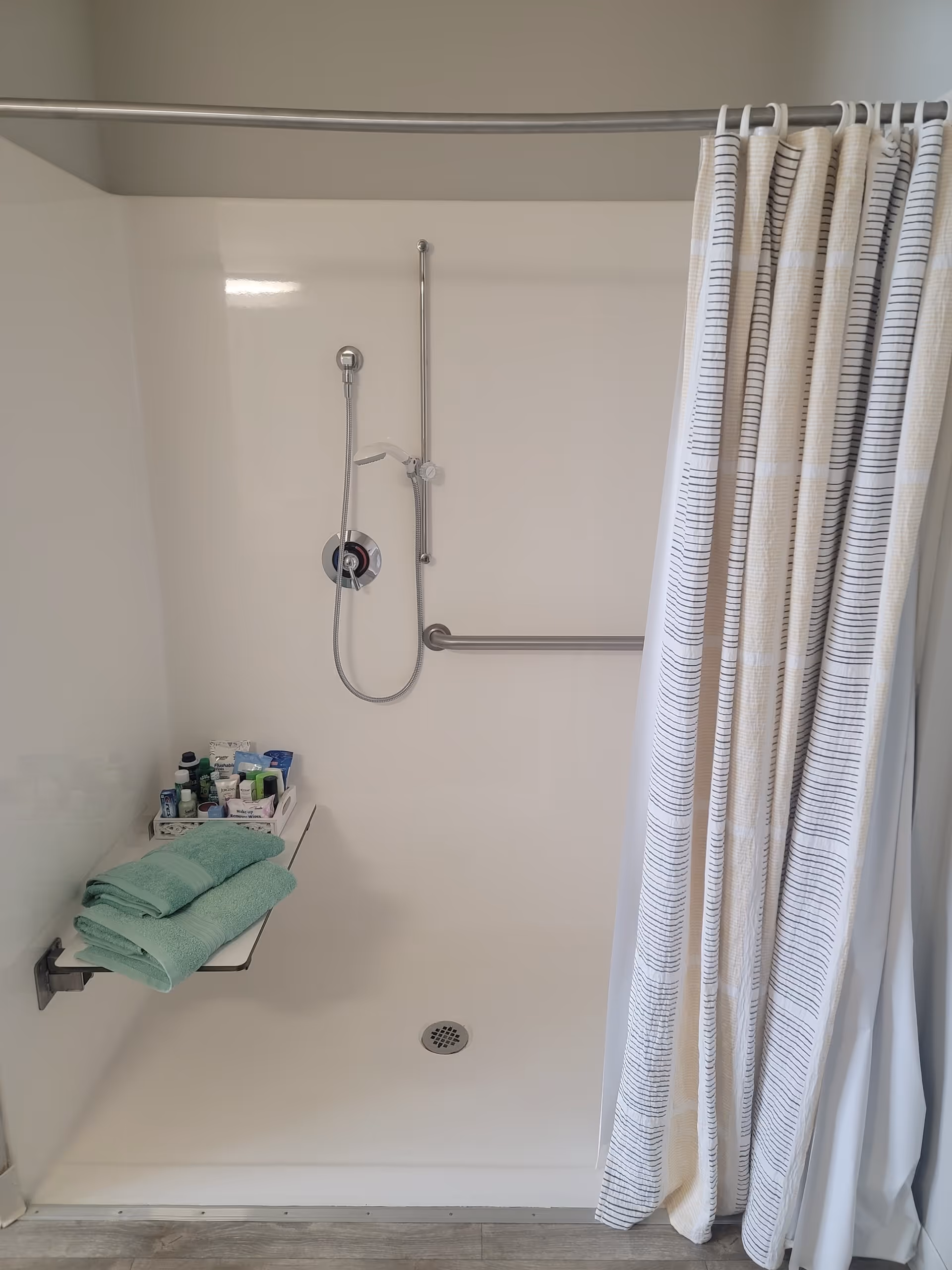 A clean, white walk-in shower with a handheld showerhead attached to a vertical bar and a horizontal grab bar on the wall. There is a fold-down shower seat holding neatly folded green towels and various toiletries. A striped shower curtain is partially drawn to the right side.
