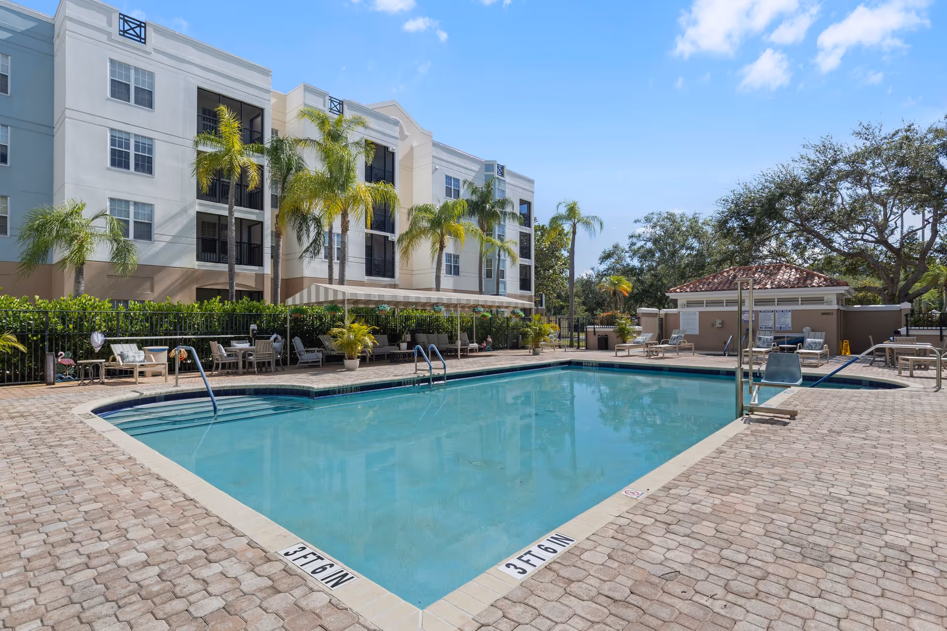 Outdoor swimming pool area with clear blue water surrounded by a paved deck. Several lounge chairs and tables with umbrellas are arranged around the pool. Palm trees and other greenery are visible along the fence line. A multi-story building with balconies is in the background under a blue sky with some clouds.