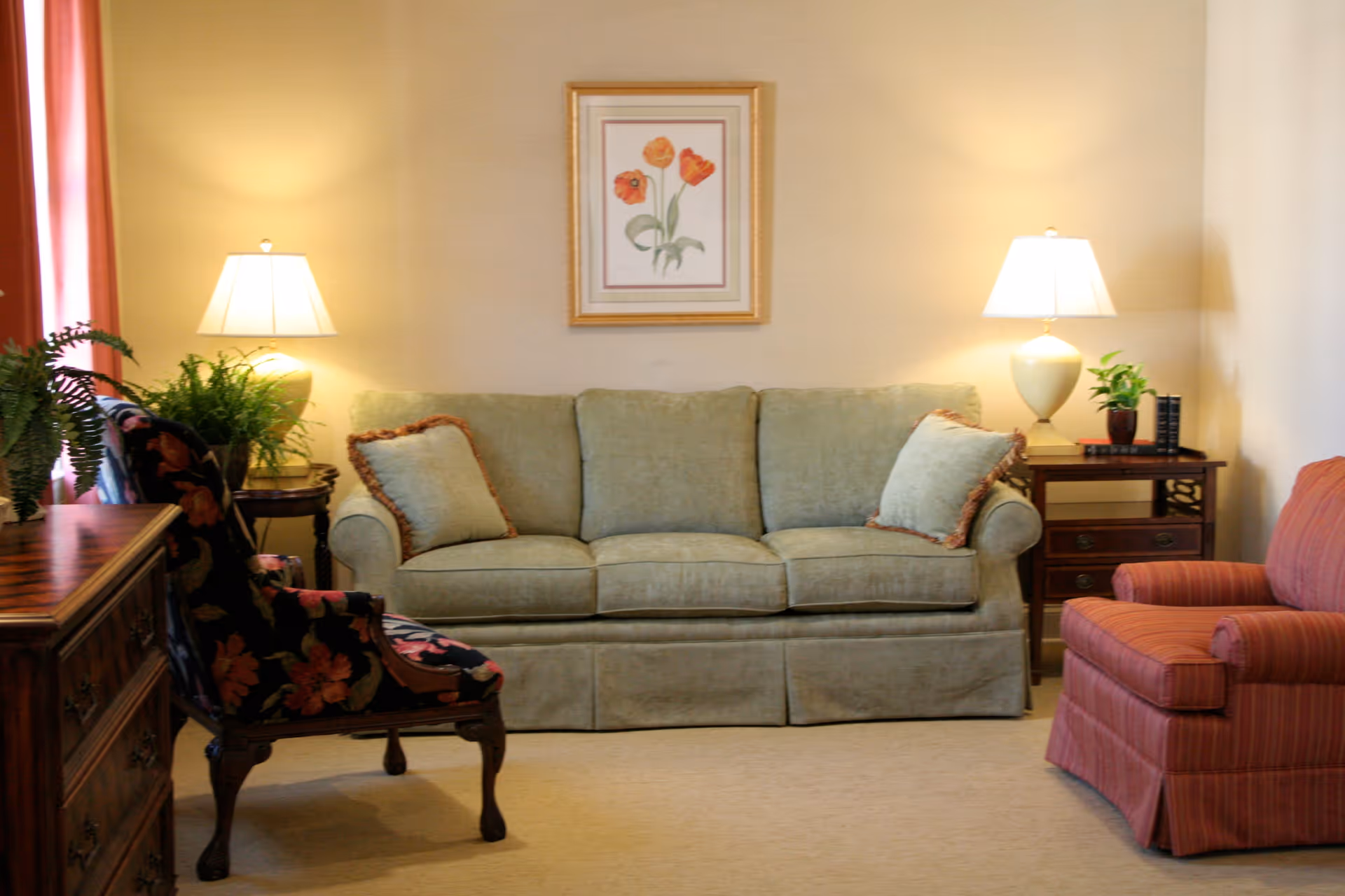 A cozy living room with a green sofa adorned with two matching pillows, a floral patterned armchair, and a red striped armchair. Two table lamps with white shades sit on wooden side tables on either side of the sofa. A framed picture of orange flowers hangs on the beige wall above the sofa. There are also green plants on the side tables and a wooden dresser on the left side of the room.