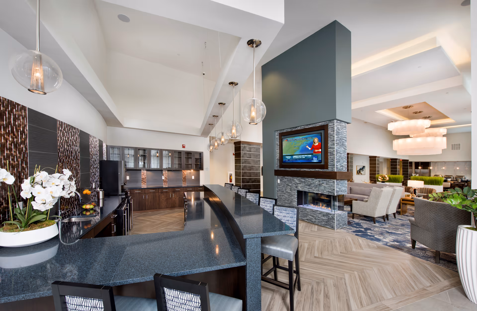 Modern communal lounge area with a long dark granite countertop bar and high chairs, pendant lights hanging from the ceiling, a fireplace with a mounted TV above it, and comfortable seating including armchairs and sofas in the background.