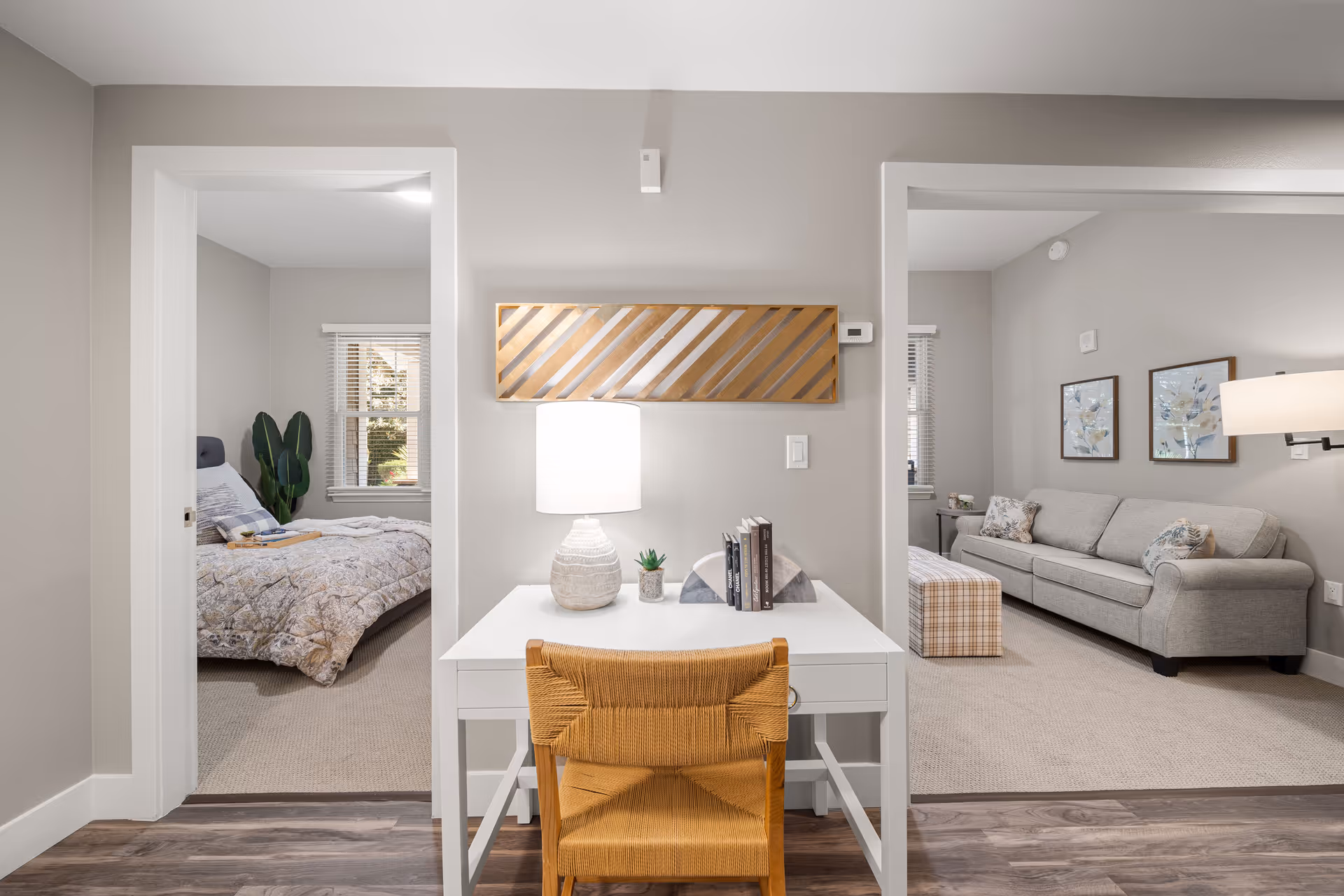 Interior view of a senior living facility showing a small white desk with a lamp, books, and a plant. To the left is a bedroom with a bed, window, and plant. To the right is a living room area with a gray sofa, two framed pictures on the wall, a plaid ottoman, and a side table with a lamp.
