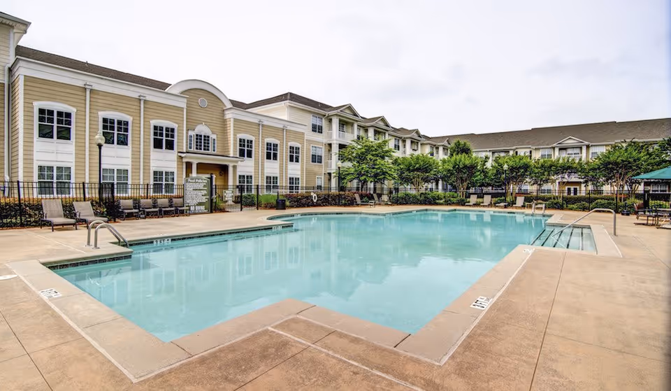 Outdoor swimming pool surrounded by a concrete deck with lounge chairs and tables. In the background, there is a multi-story residential building with beige siding and white trim, along with trees and greenery around the pool area.