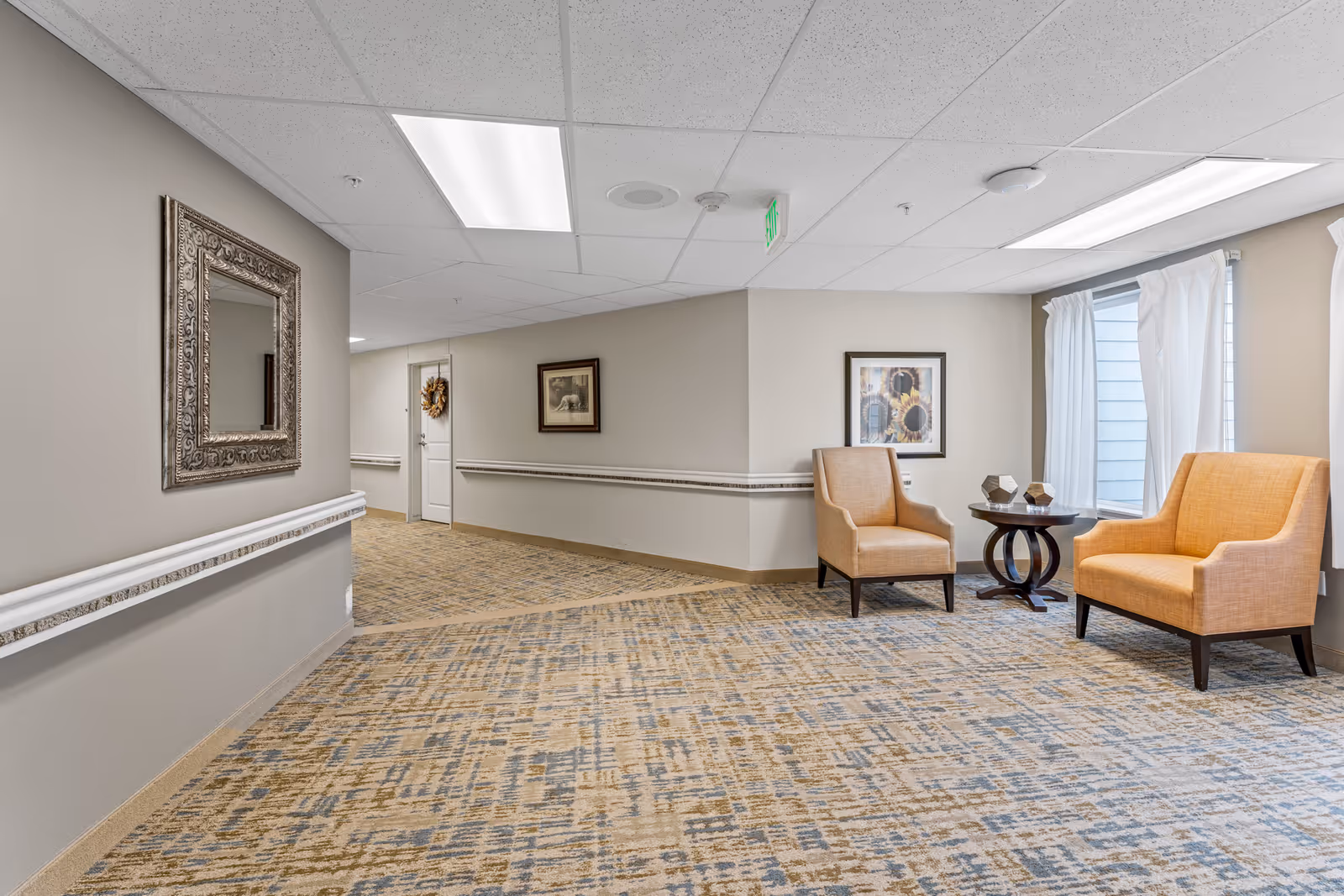 A carpeted senior living facility hallway with two upholstered chairs and a small table beside a window.