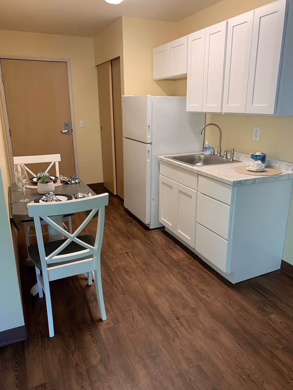 Small kitchenette with white cabinets, refrigerator, sink, and a two-person dining table set on wood flooring.