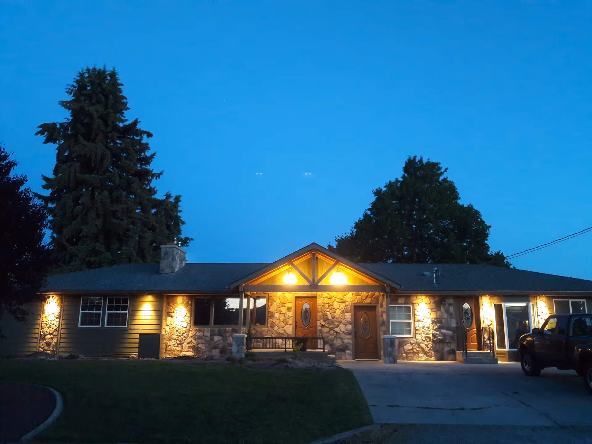 Exterior view of a single-story building at dusk with warm lights illuminating the stone and wood facade. The building has multiple doors and windows, a small covered porch area with a bench, and is surrounded by trees and a lawn. A black pickup truck is parked on the driveway to the right.
