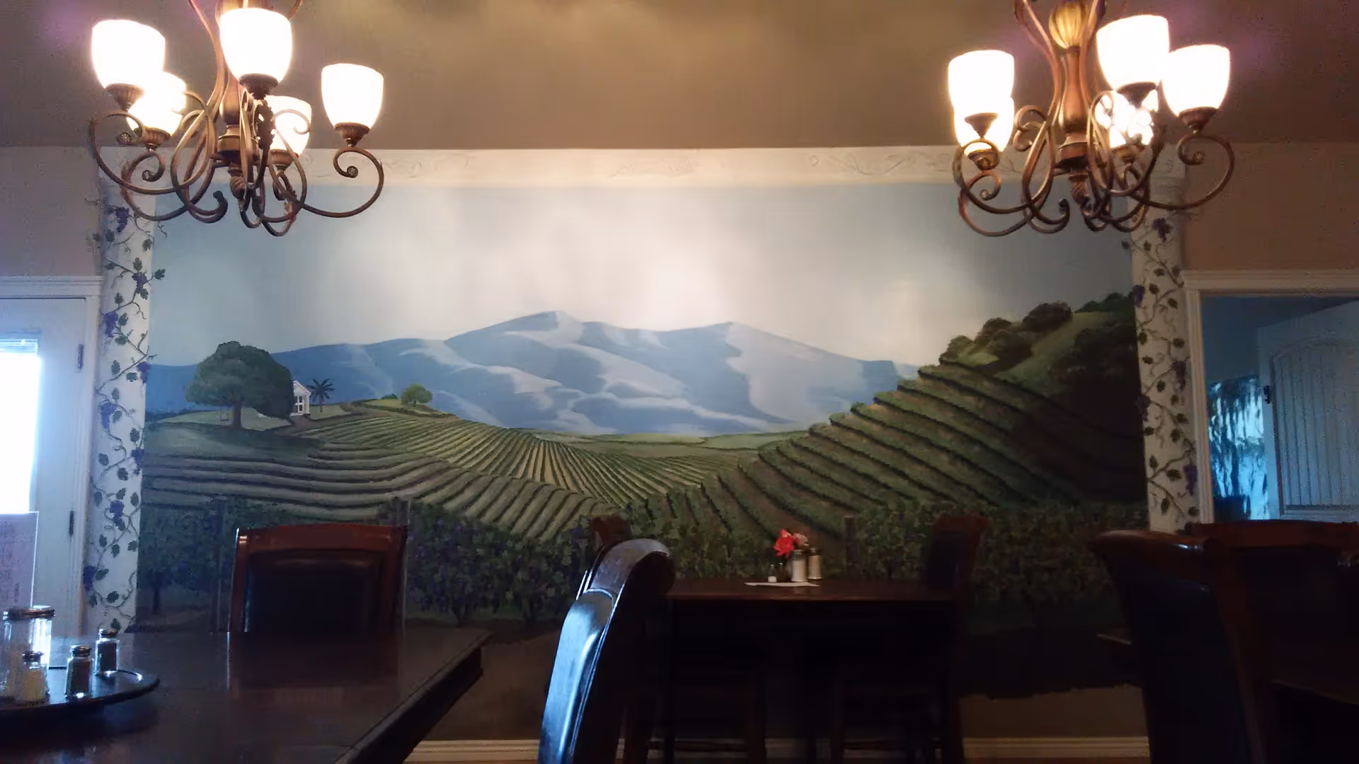 Dining room with a large vineyard mural on the wall, wooden tables and chairs, and two chandeliers overhead.