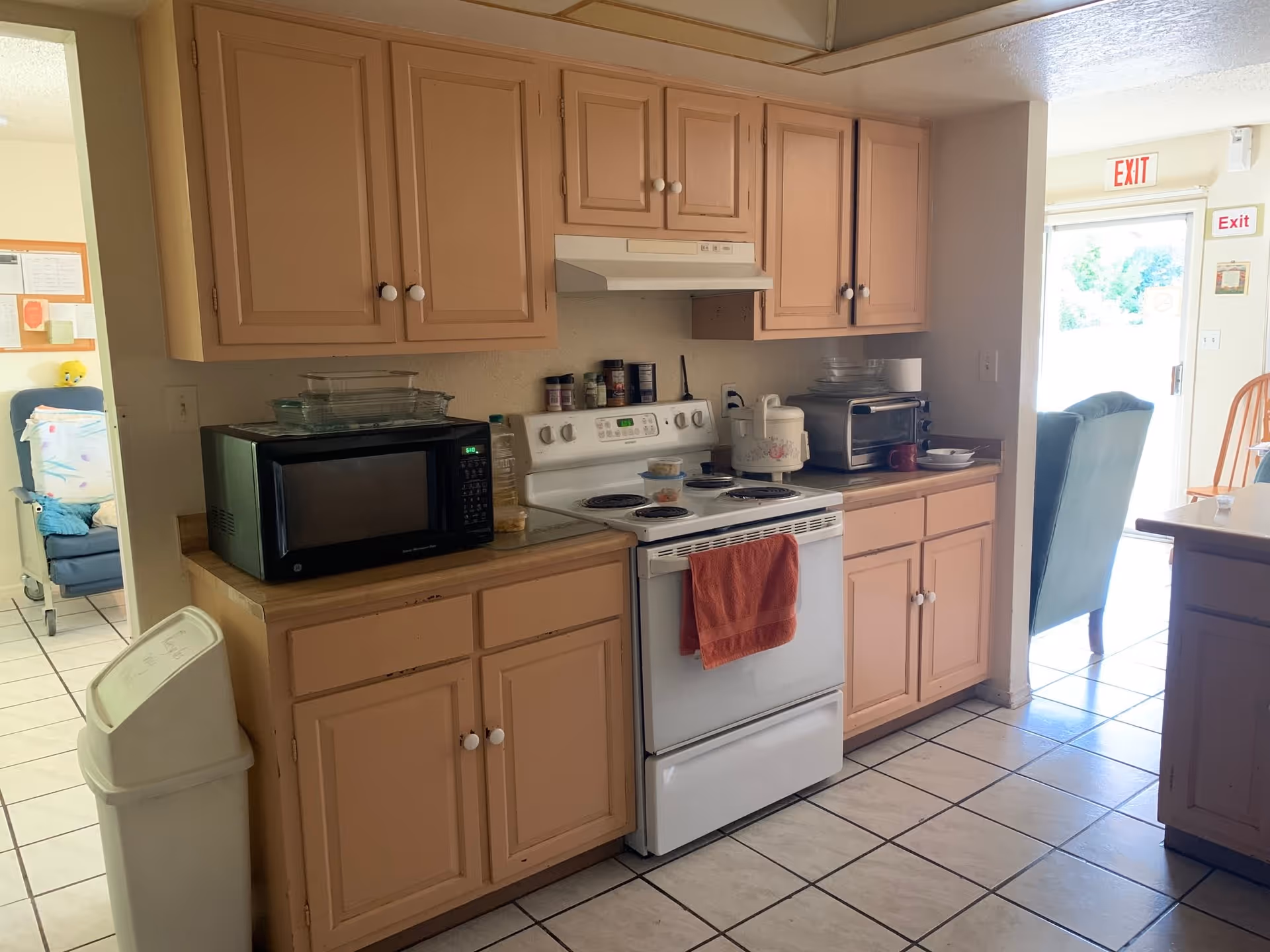 A kitchen area in a senior living facility with beige cabinets, a white electric stove with a red towel hanging on the handle, a black microwave on the counter, a toaster oven, and various kitchen items on the countertops. There is a trash can to the left and a doorway leading to a room with a blue armchair and a window letting in natural light.
