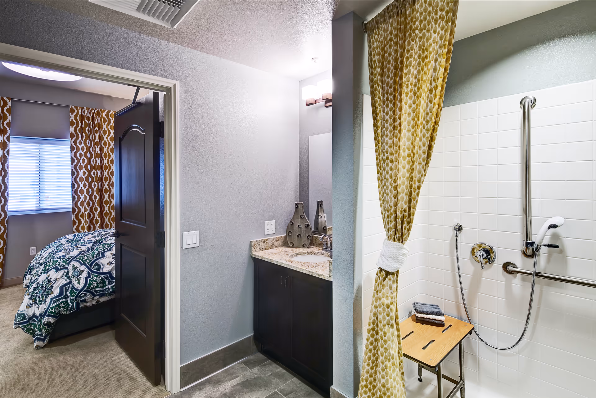 Accessible bathroom with a walk-in tiled shower with grab bars and a patterned shower curtain, a vanity, and a view into a bedroom.