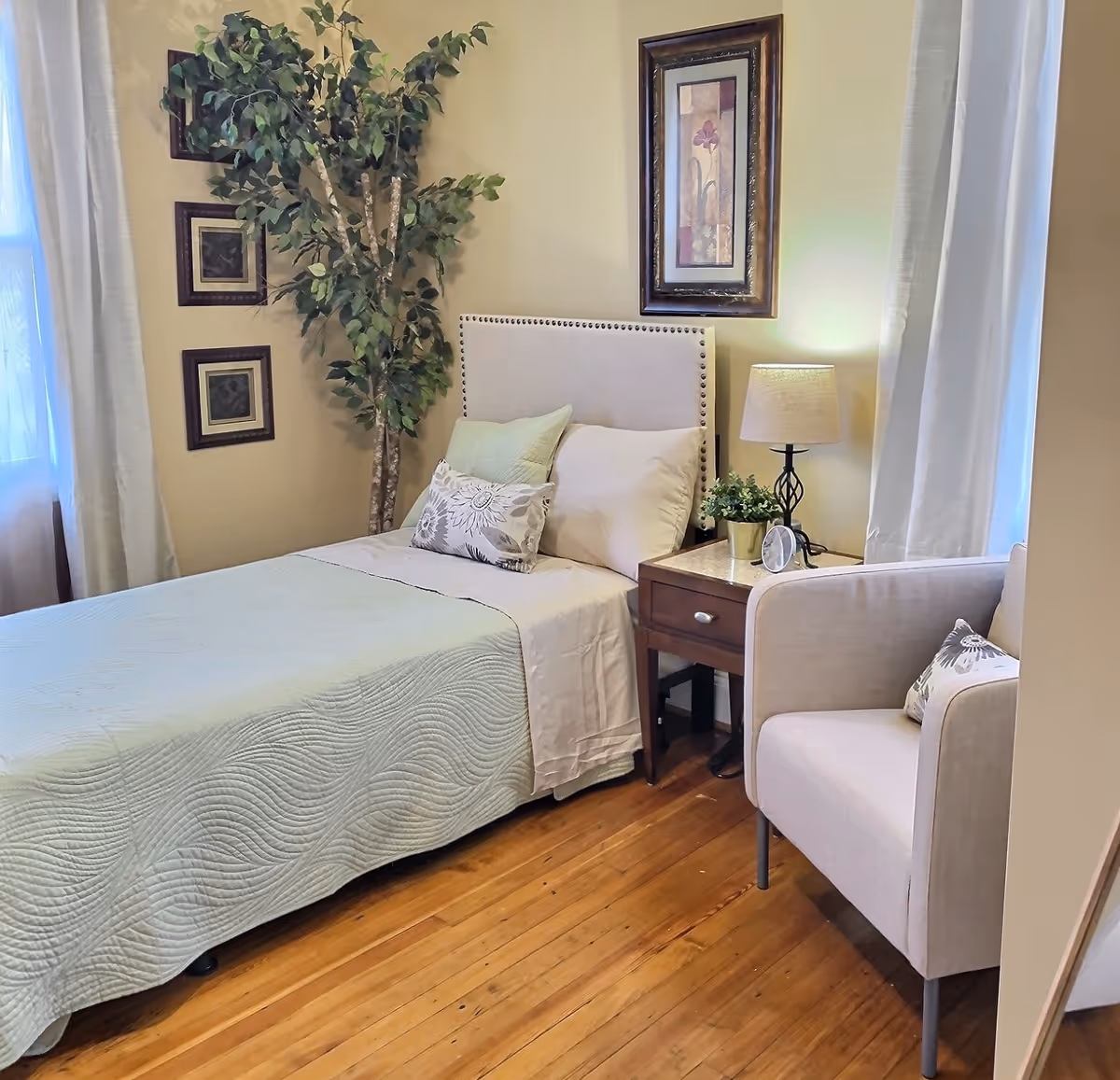 A cozy bedroom with a single bed covered in a light green quilt and decorative pillows. Next to the bed is a wooden nightstand with a lamp, a small plant, and a clock. A beige armchair with a patterned pillow is positioned near the nightstand. The room has wooden flooring, light-colored walls, two windows with white curtains, and framed artwork on the walls. A tall artificial plant is placed in the corner.