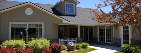 Exterior view of a single-story residential building with beige siding, large windows, a small white picket fence, and a landscaped front yard with bushes and a tree with autumn leaves under a clear blue sky.