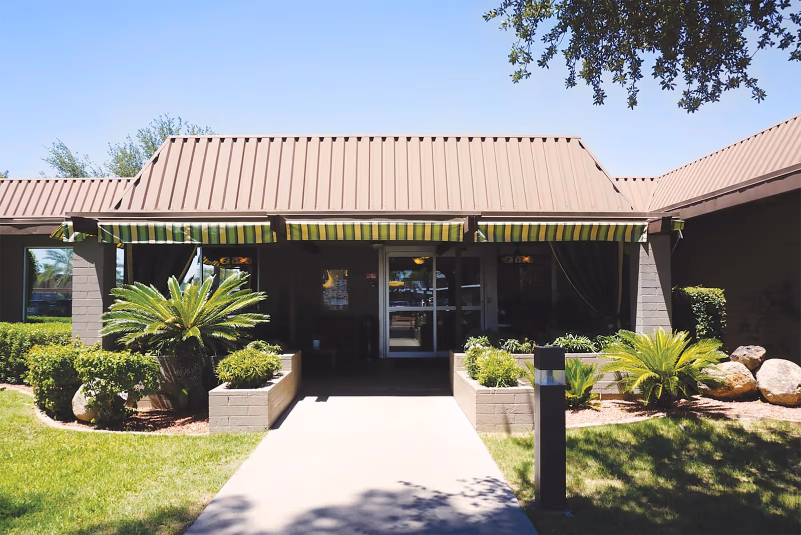 Front exterior view of Haven Health Phoenix facility with a brown metal roof, striped green and yellow awnings, a concrete walkway leading to glass entrance doors, and landscaped greenery including bushes and small palm plants.