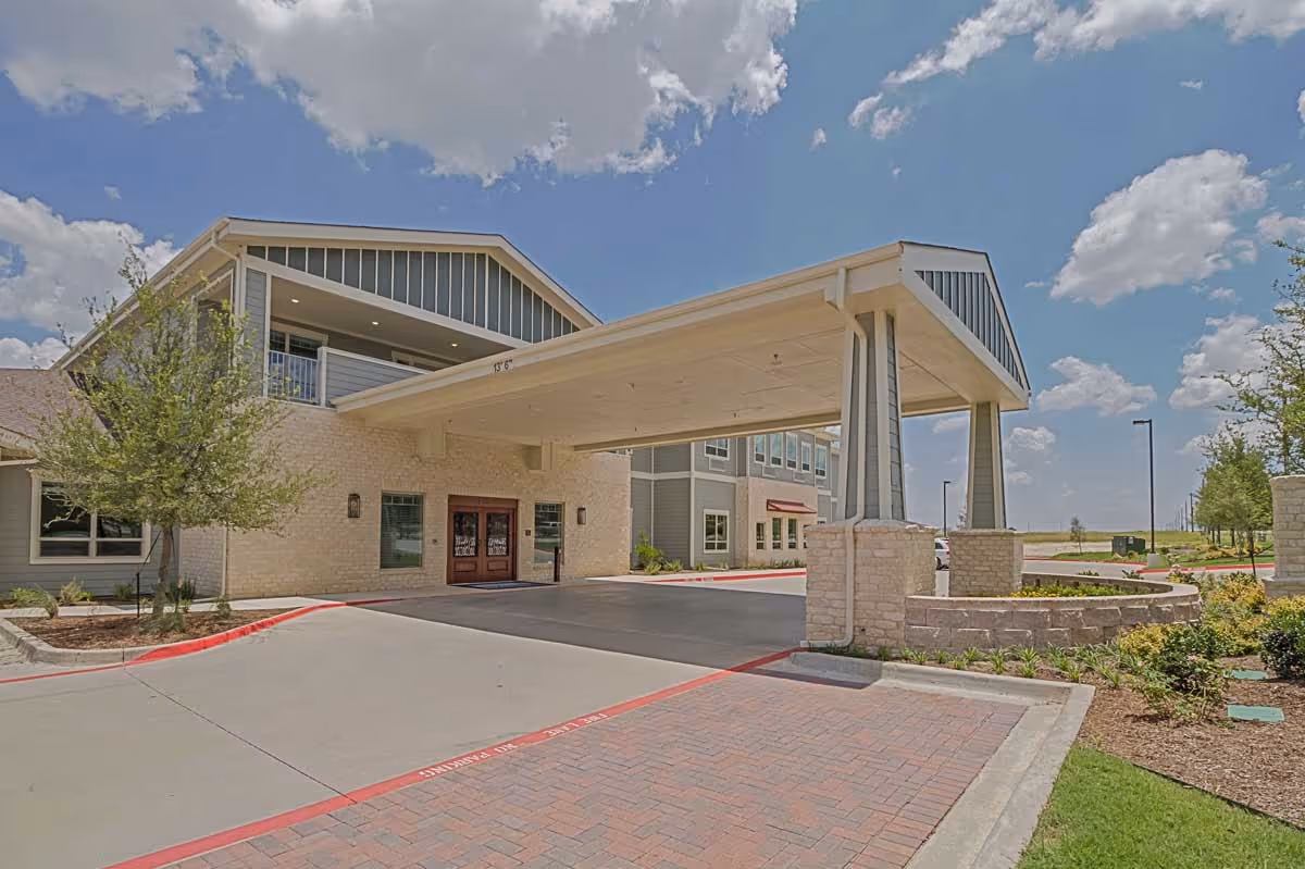 Exterior view of Midtowne Assisted Living and Memory Care building entrance with a covered drop-off area, stone and siding facade, landscaped surroundings, and a partly cloudy sky.