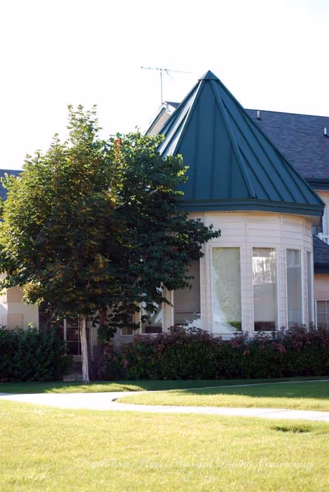 Front exterior of a light-colored building with a green conical roof, large windows, a tree, and landscaped lawn.