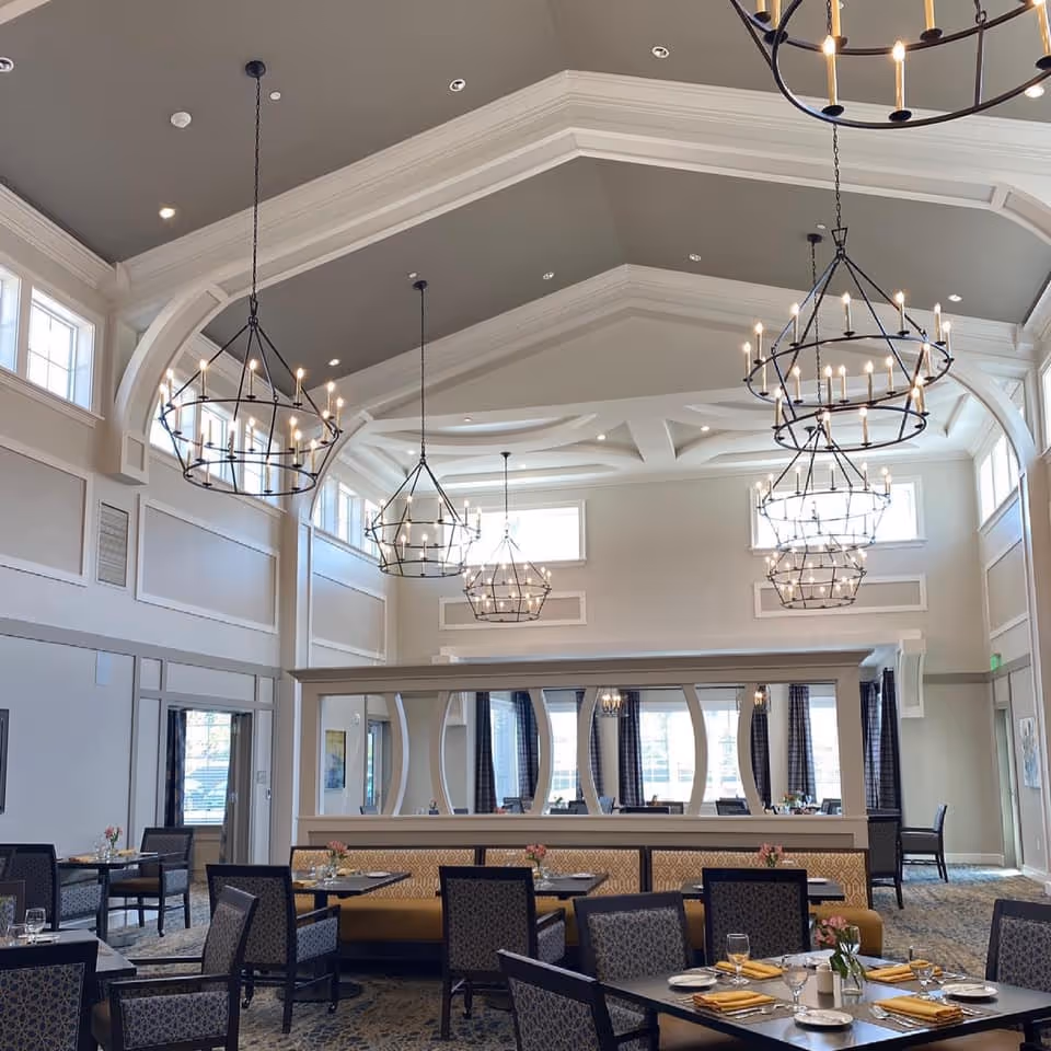 A spacious dining room with high vaulted ceilings and multiple large, circular chandeliers hanging from the ceiling. The room features several dining tables with chairs, each set with plates, glasses, and yellow napkins. There is a decorative partition with mirrors in the middle of the room, and large windows near the ceiling allow natural light to fill the space.