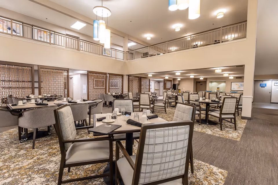 A spacious dining room in a senior living facility with multiple tables and chairs arranged neatly. The room features a high ceiling with modern hanging light fixtures, patterned carpet, and decorative wooden lattice panels along one wall. The tables are set with glasses, napkins, and menus.