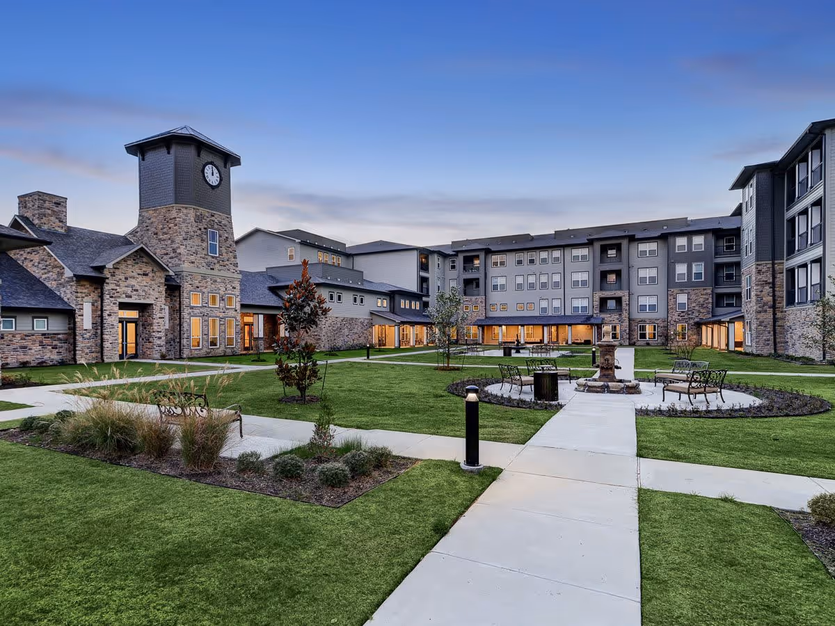 Exterior view of The Lodge at Pine Creek senior living facility during dusk, showing a large courtyard with green lawns, paved walkways, benches, and a central fountain. The building features stone and gray siding with multiple windows and a clock tower.