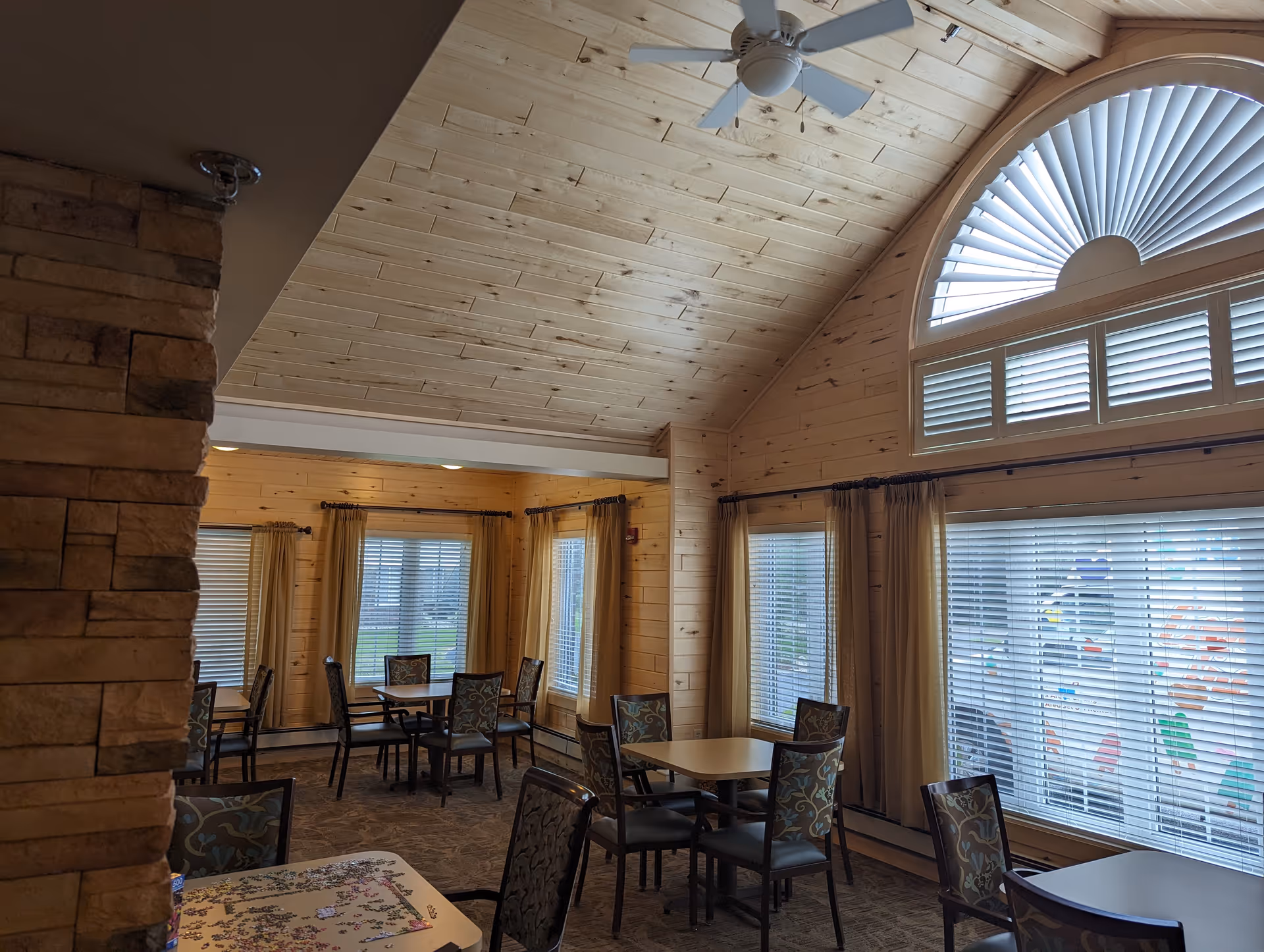 Interior view of a dining area with multiple tables and chairs. The room features wooden walls and a high, slanted wooden ceiling with a ceiling fan. Large windows with blinds and curtains allow natural light to enter the space.