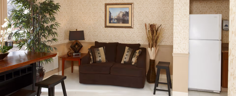 A cozy living area featuring a dark brown loveseat with patterned cushions, a wooden side table with a lamp, a tall vase with decorative dried plants, and a white refrigerator partially visible in an adjacent kitchen space. There is a framed painting on the wall above the loveseat, a large potted plant near a window, and two black stools near a wooden counter.