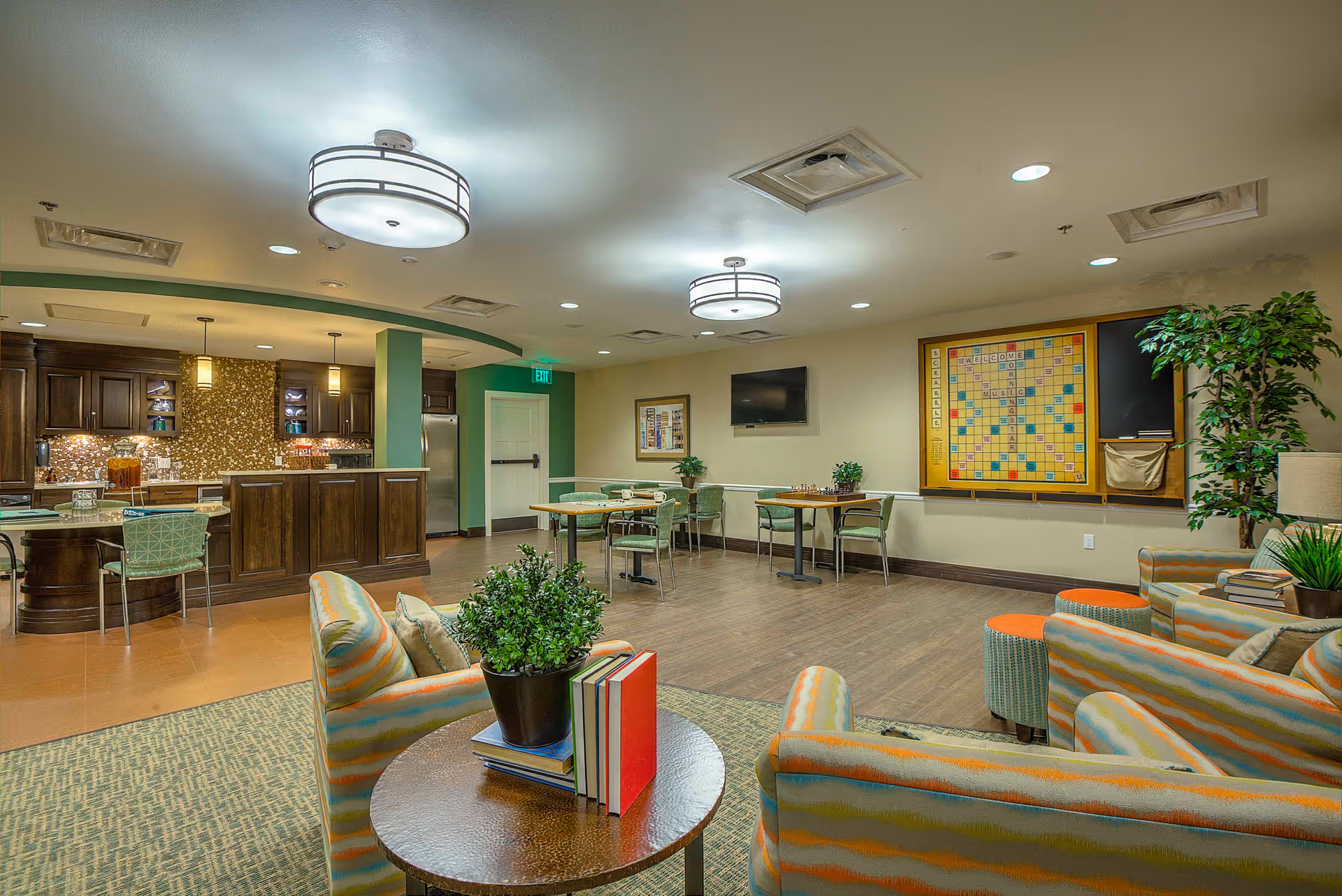 A spacious and well-lit common area in an assisted living facility featuring a seating area with patterned armchairs and a round table with books and a potted plant. In the background, there is a kitchen area with dark wood cabinets and a countertop with chairs. On the far wall, there are tables and chairs, a large Scrabble board mounted on the wall, a flat-screen TV, and some potted plants. The ceiling has modern light fixtures and recessed lighting.