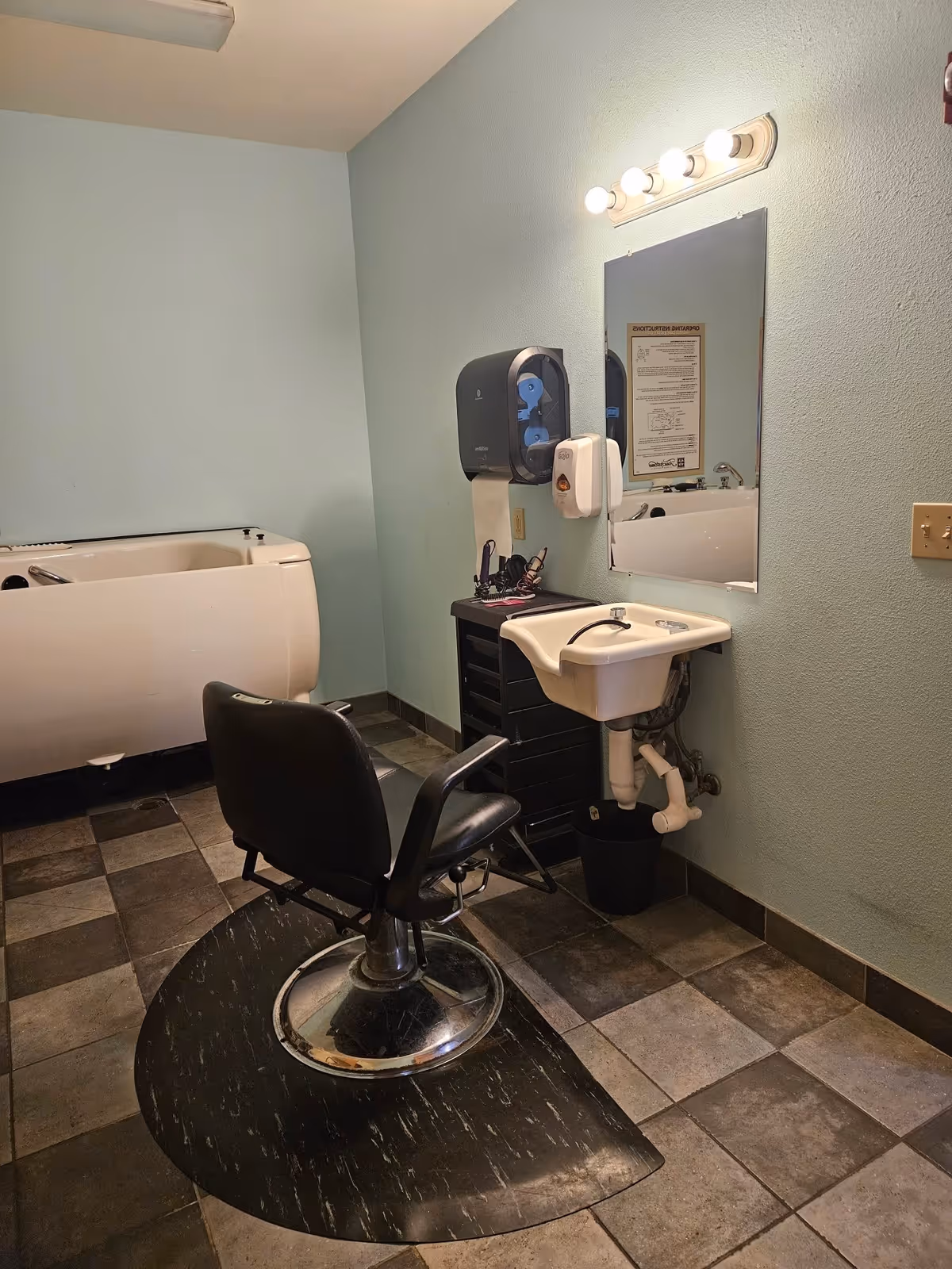 A small salon area inside a facility with a black salon chair on a black mat, a white sink attached to the wall, a mirror above the sink, a paper towel dispenser, and a hair washing station in the background. The walls are painted light blue and the floor is tiled with a mix of dark and light brown tiles.