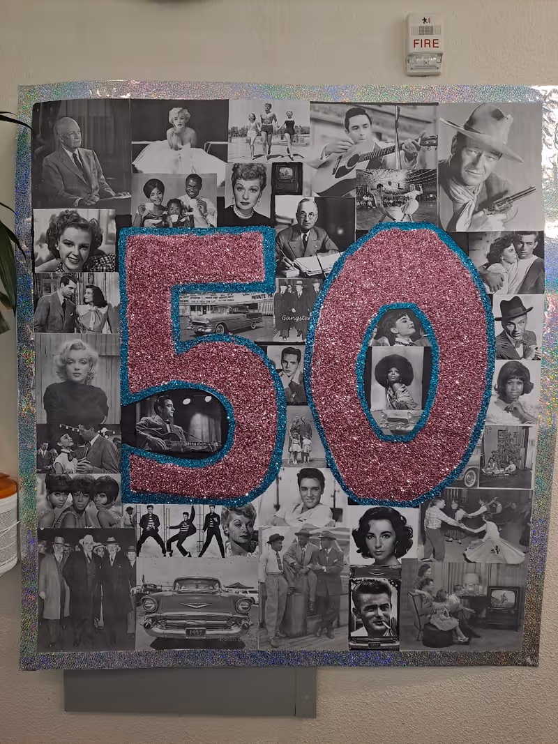 Decorative collage of black-and-white vintage photos with a large glittery pink '50' outlined in blue mounted on a wall beneath a fire alarm.