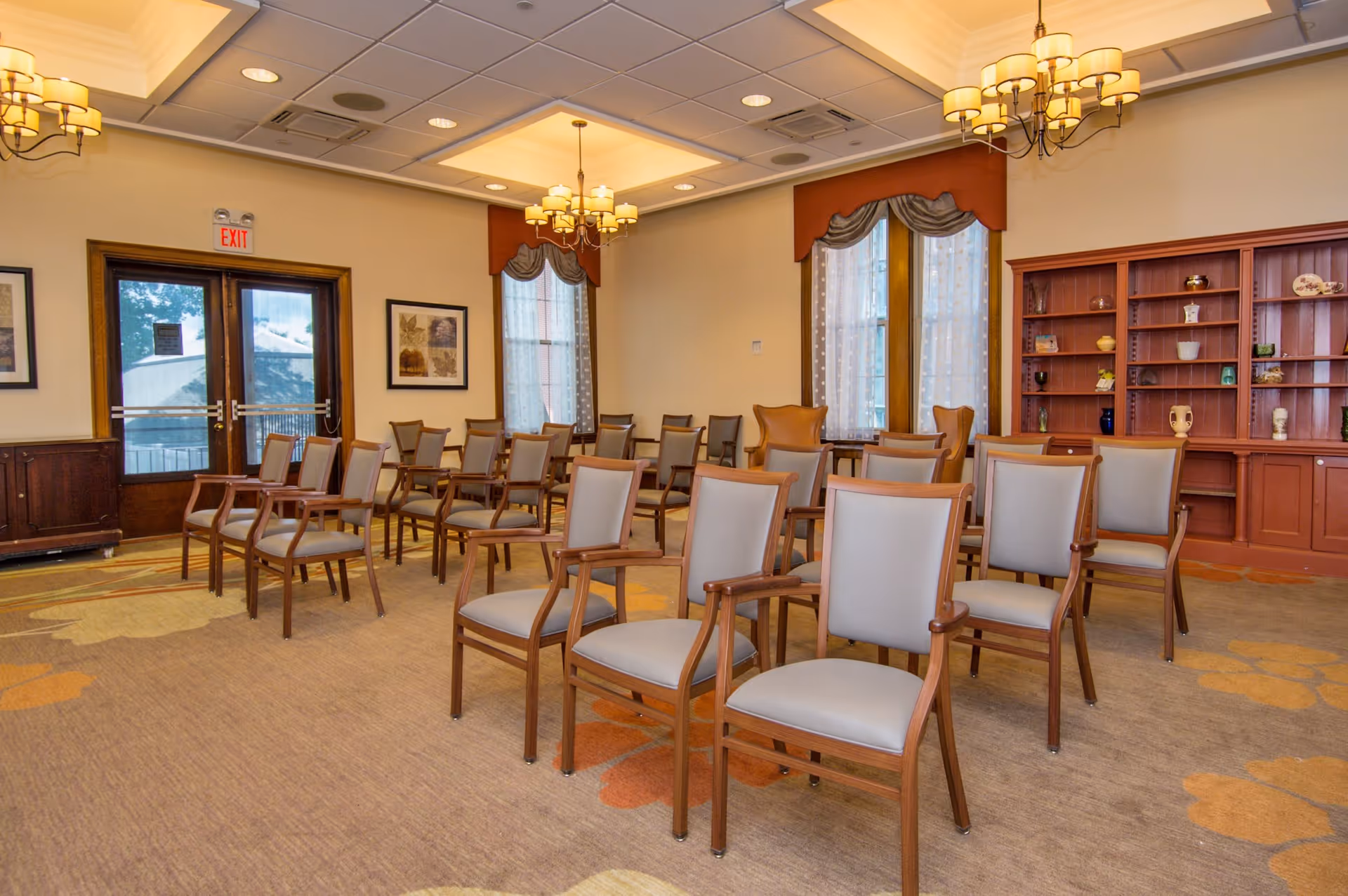 A well-lit room with multiple wooden chairs arranged in rows facing the front, featuring beige cushioned seats and backs. The room has large windows with sheer curtains and valances, two chandeliers hanging from the ceiling, framed artwork on the walls, and a wooden built-in bookshelf filled with decorative items. Double glass doors with an exit sign above are visible on one side.
