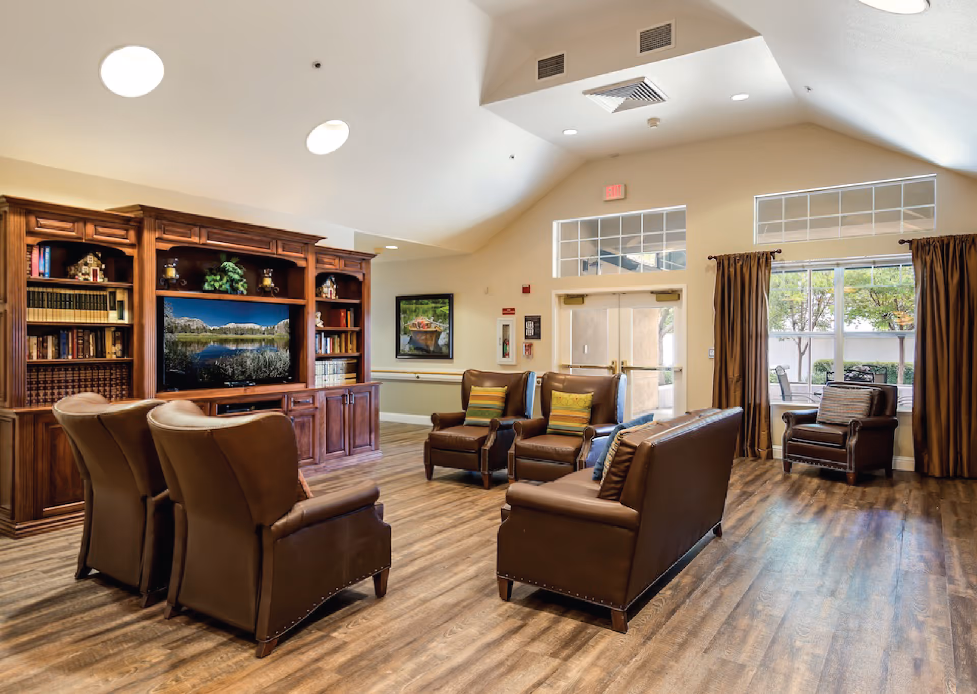 A spacious senior living facility common area with wooden flooring, brown leather chairs and sofas arranged around a large wooden entertainment center with a TV and bookshelves. The room has high ceilings with recessed lighting and large windows with brown curtains letting in natural light. There is a door leading outside to a patio area with outdoor furniture.