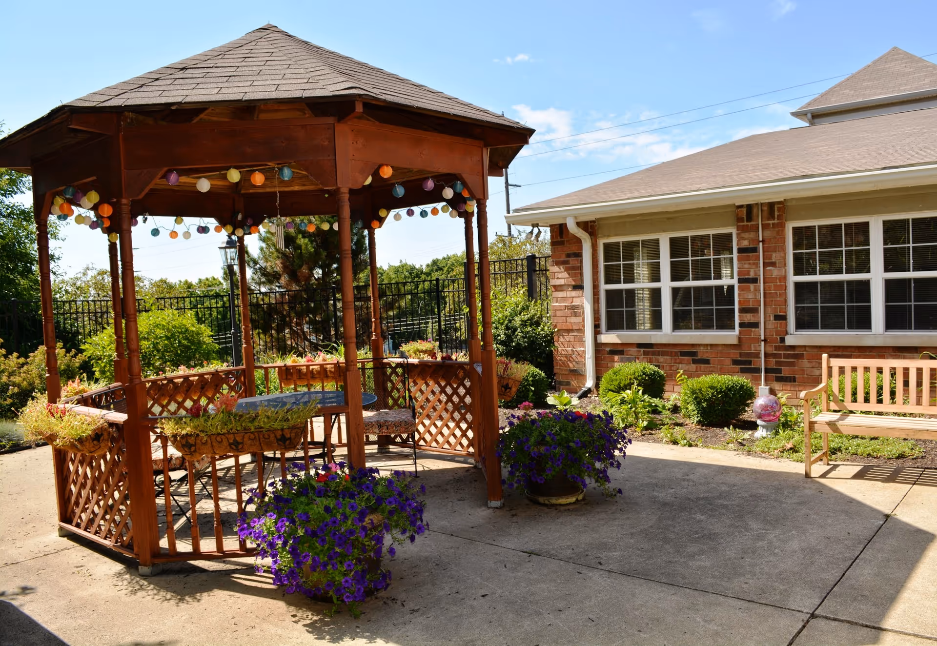 Outdoor patio area at Rosewalk Village of Lafayette featuring a wooden gazebo decorated with colorful string lights, surrounded by potted flowers and greenery. There is a wooden bench near a brick building with large windows under a clear blue sky.