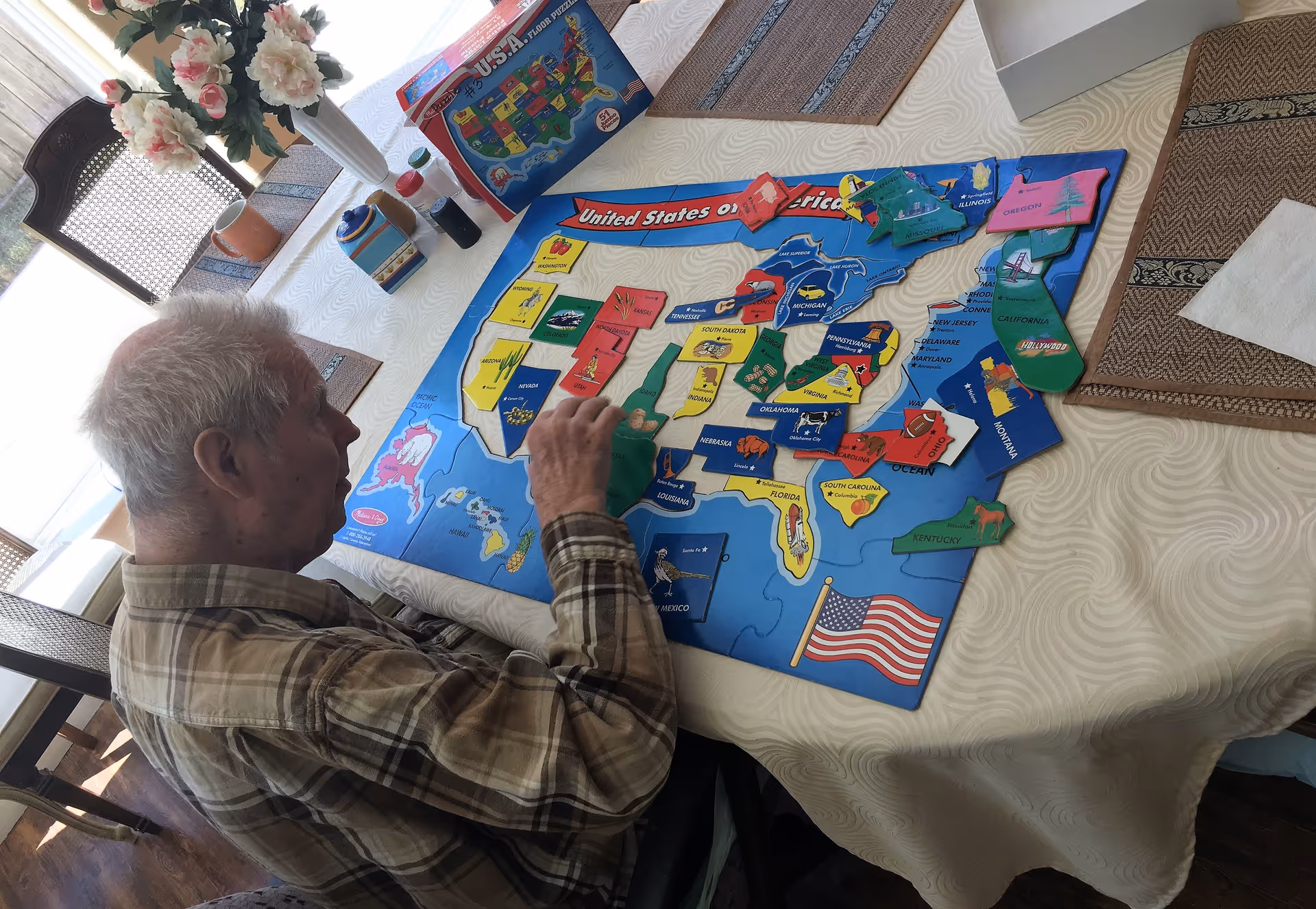 An elderly man sitting at a table working on a colorful wooden puzzle map of the United States. The table is covered with a white tablecloth and has a vase with flowers and some other items on it. The room is well-lit with natural light coming from a window.