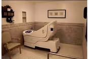 A walk-in bathtub with a built-in seat in a tiled senior bathroom, with a chair and grab bars nearby.