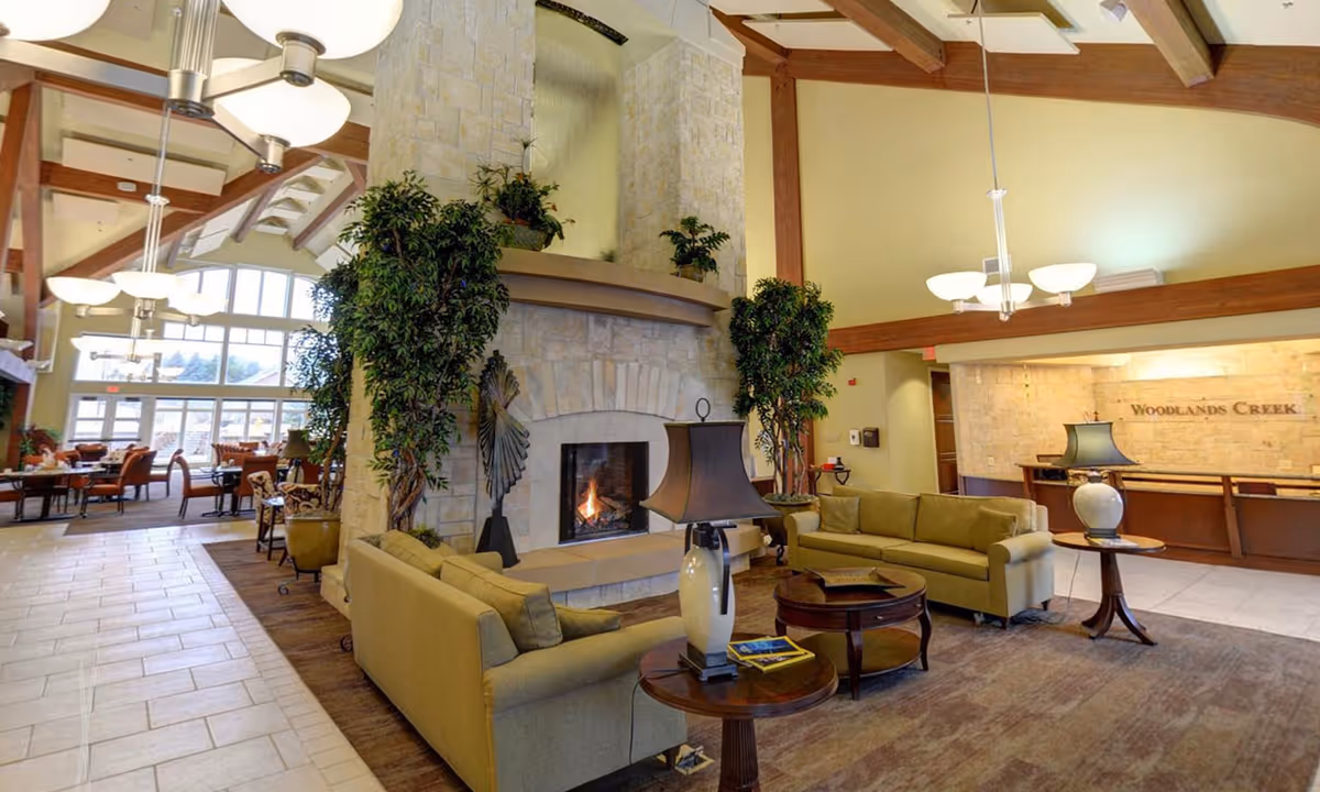 Spacious senior living lobby/lounge with sofas, a stone fireplace, tall windows, and a reception desk labeled 'Woodlands Creek'.
