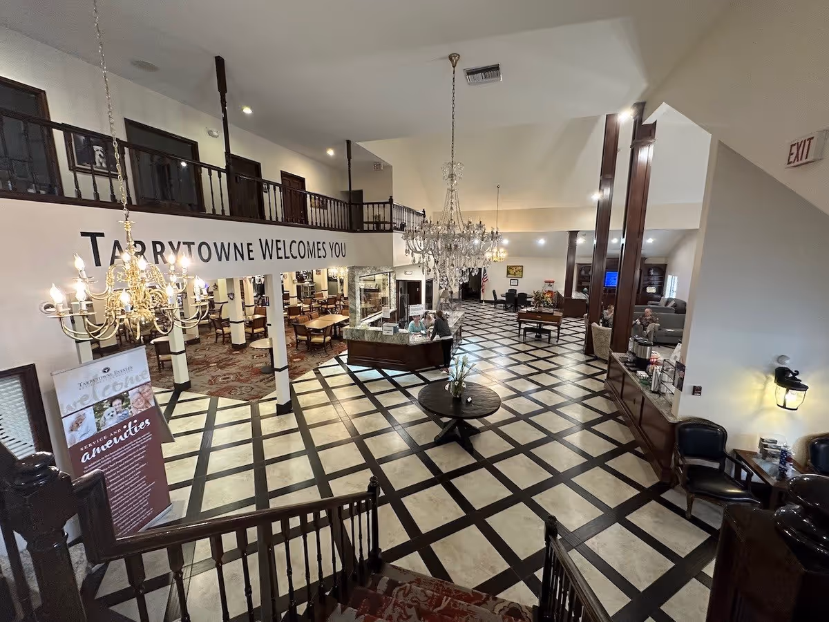 Interior view of Tarrytowne Estates featuring a spacious lobby with checkered tile flooring, a round table with a flower arrangement in the center, chandeliers hanging from the ceiling, a reception desk with two staff members, seating areas with chairs and sofas, and a dining area visible in the background. The wall has a sign that reads 'TARRYTOWNE WELCOMES YOU'.
