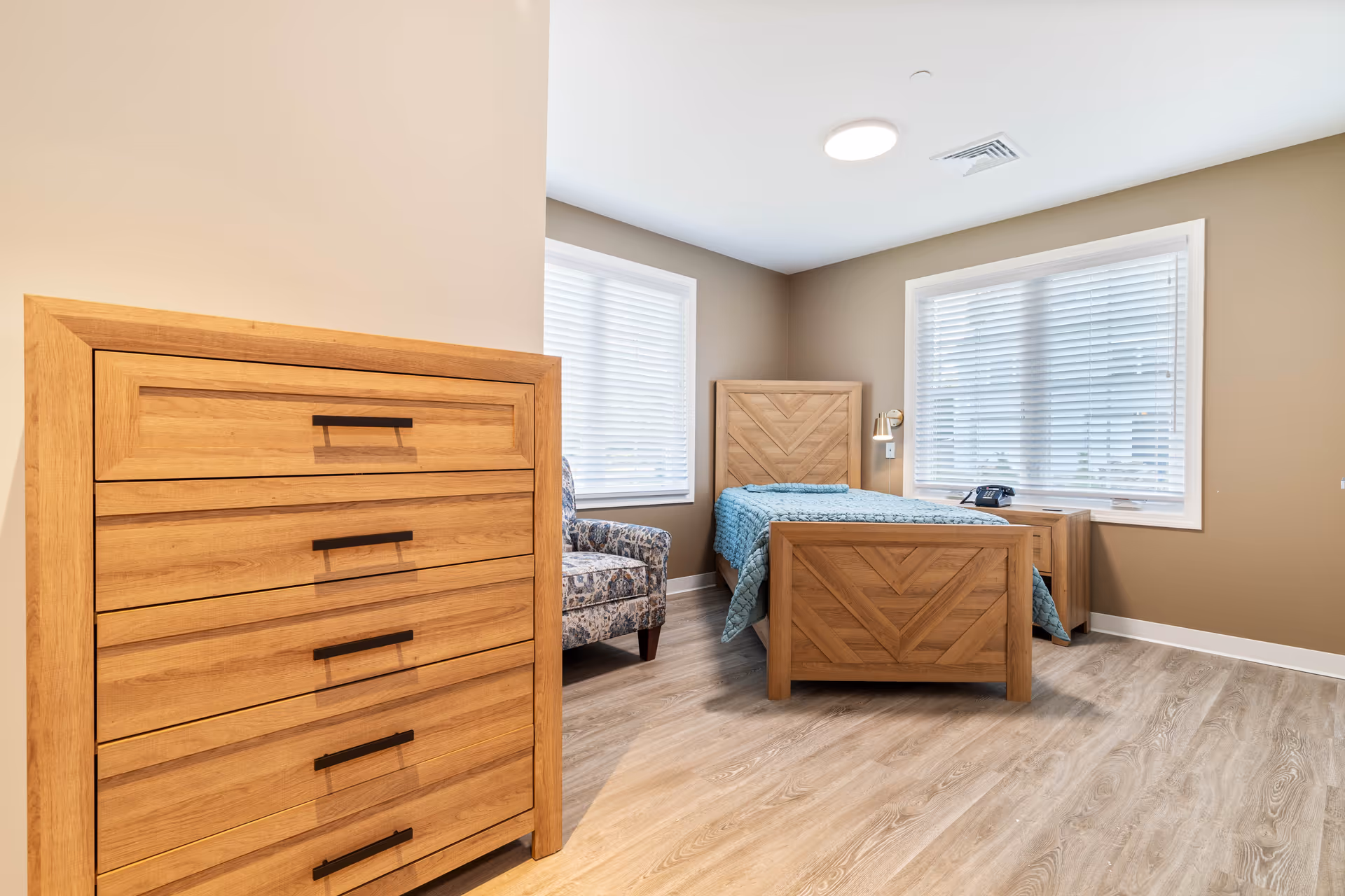 A bright and clean bedroom with light brown walls and wood flooring. The room features a wooden bed with a blue quilt, a matching wooden dresser with five drawers, a wooden nightstand with a black telephone, and a patterned armchair. Two windows with white blinds allow natural light to fill the room.