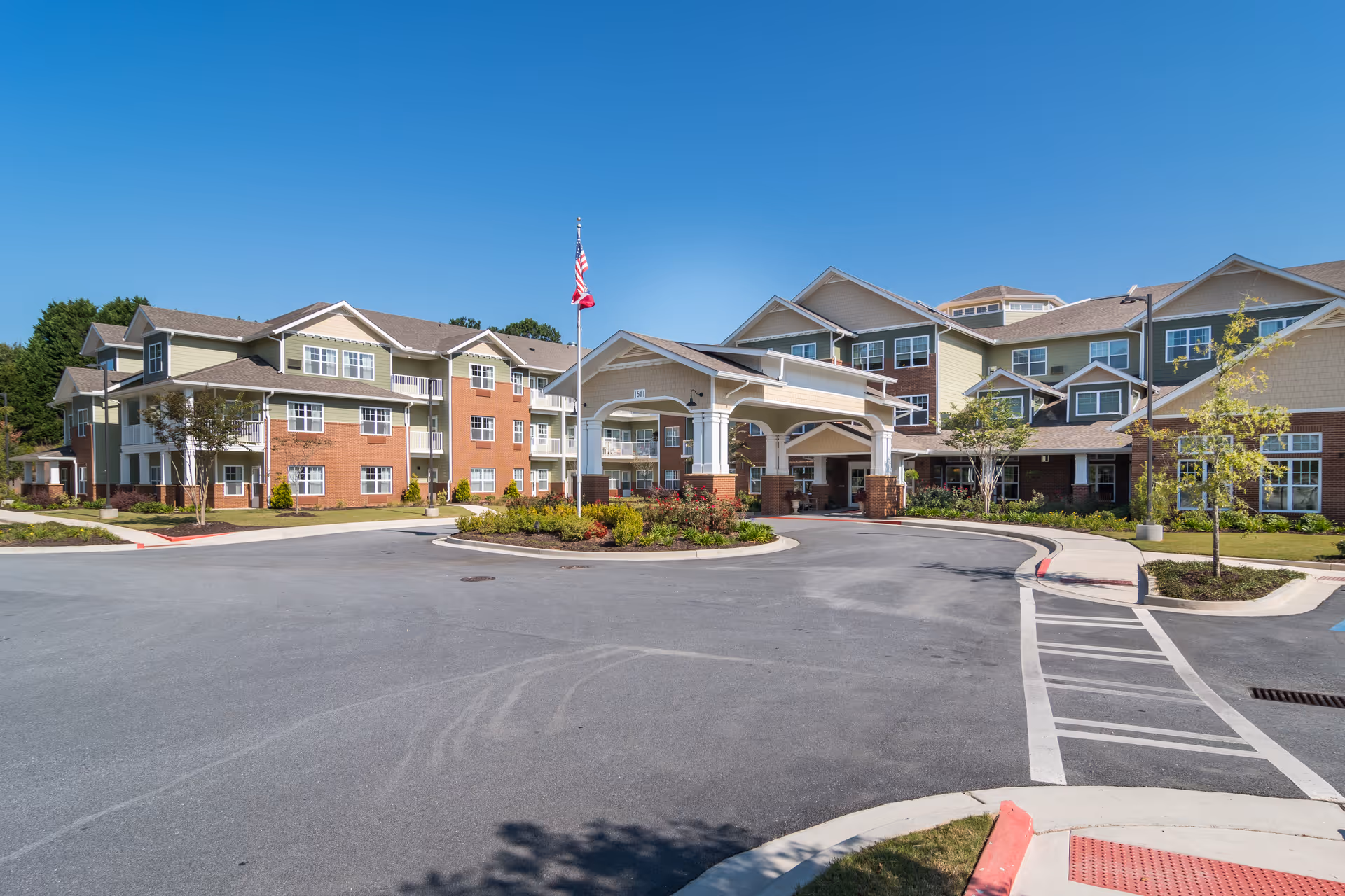 Exterior view of Linwood Estates Gracious Retirement Living facility showing a large multi-story building with a covered entrance, landscaped roundabout, and an American flag on a flagpole under a clear blue sky.