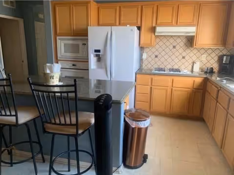 Kitchen with a center island and two bar stools, light wood cabinets, white refrigerator and built-in microwave, and a tiled backsplash.